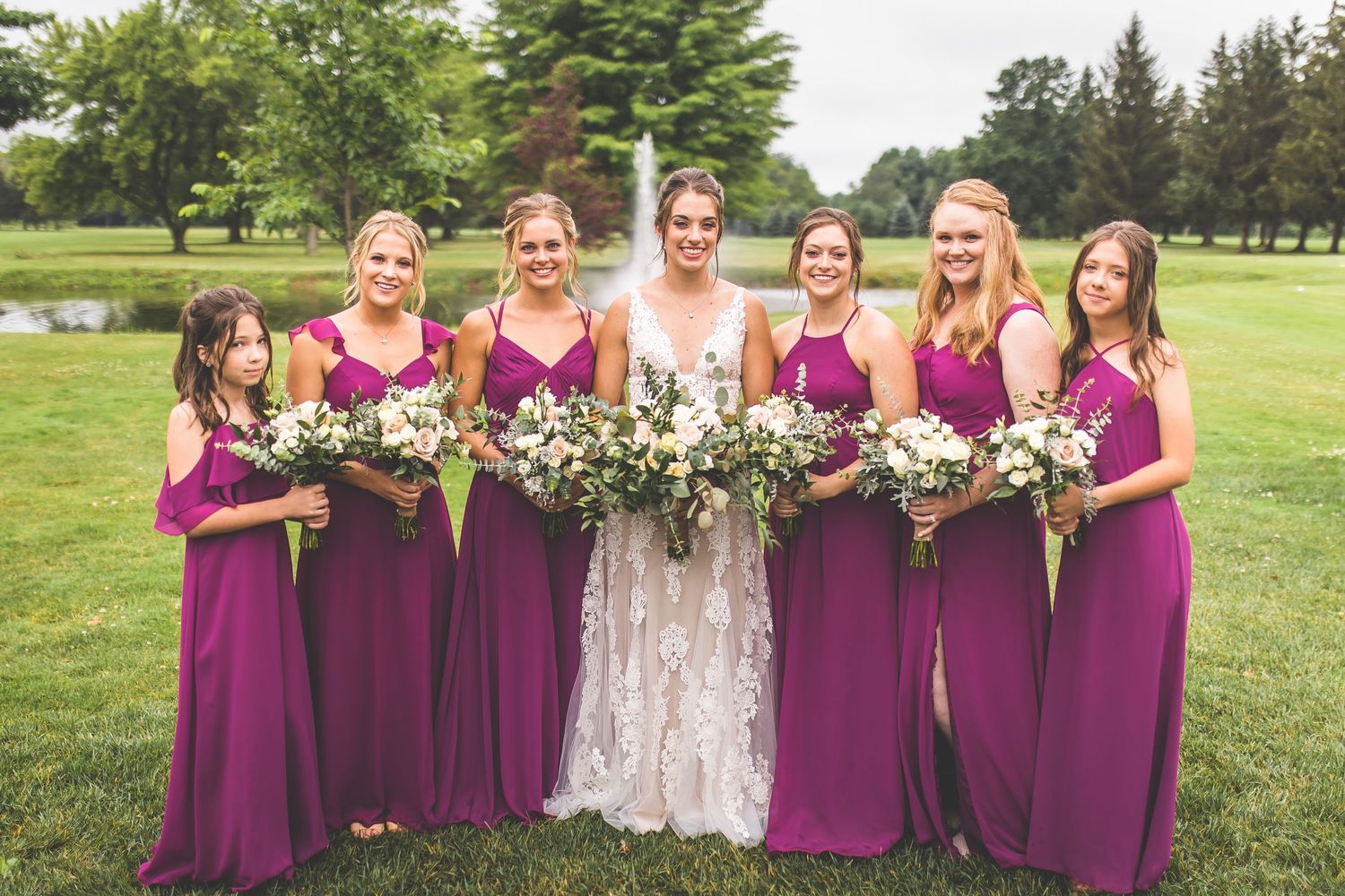 Bridesmaids in burgundy dresses pose with the bride holding floral bouquets on a sunny lawn.