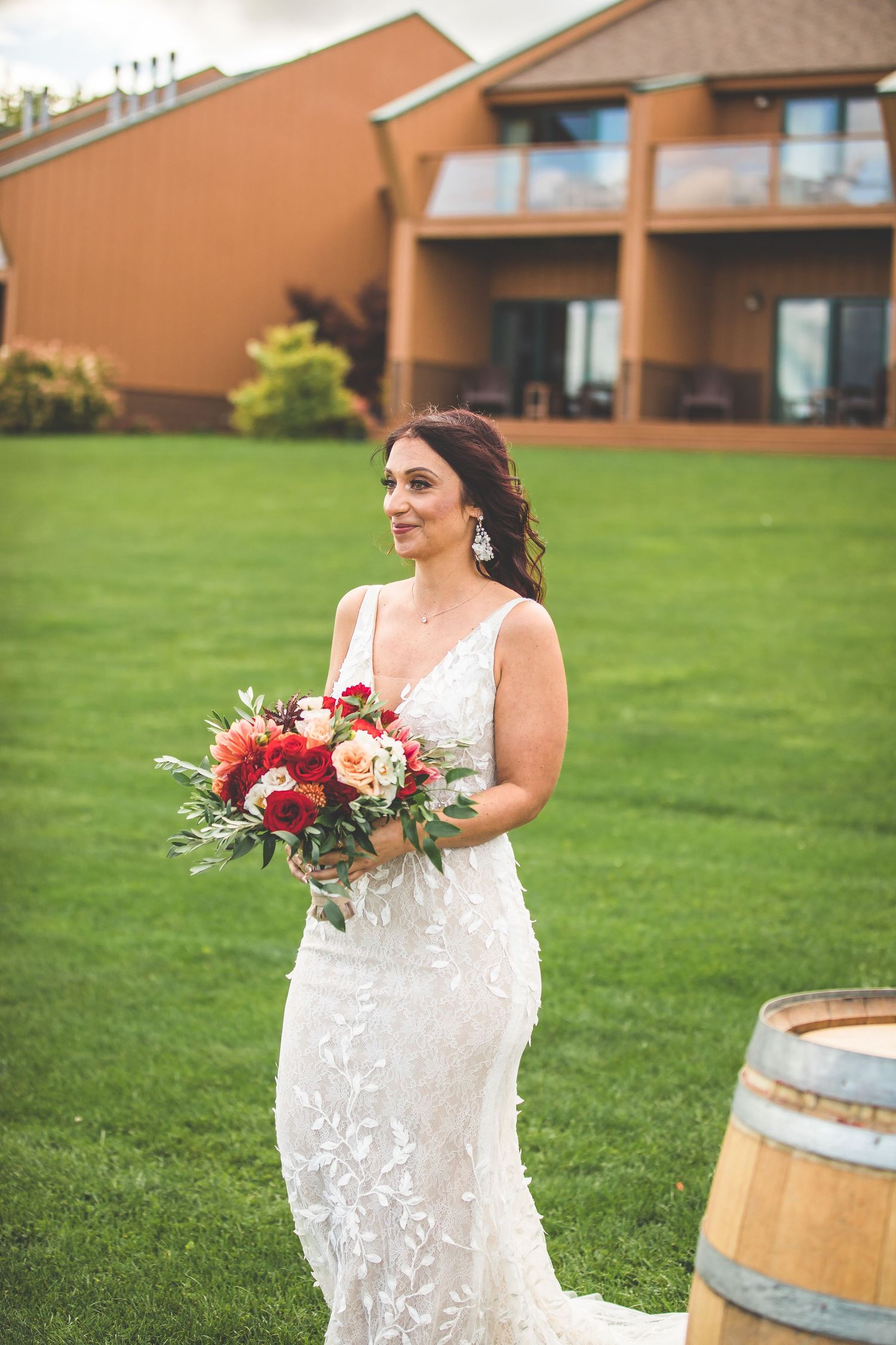 Bride in lace wedding dress holds red and peach bouquet near rustic wine barrel outdoors.