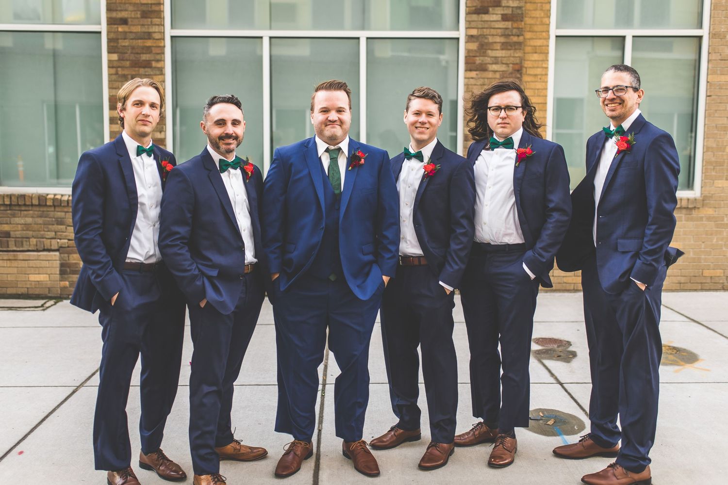 Groomsmen in navy suits and red boutonnieres pose together against a brick building.