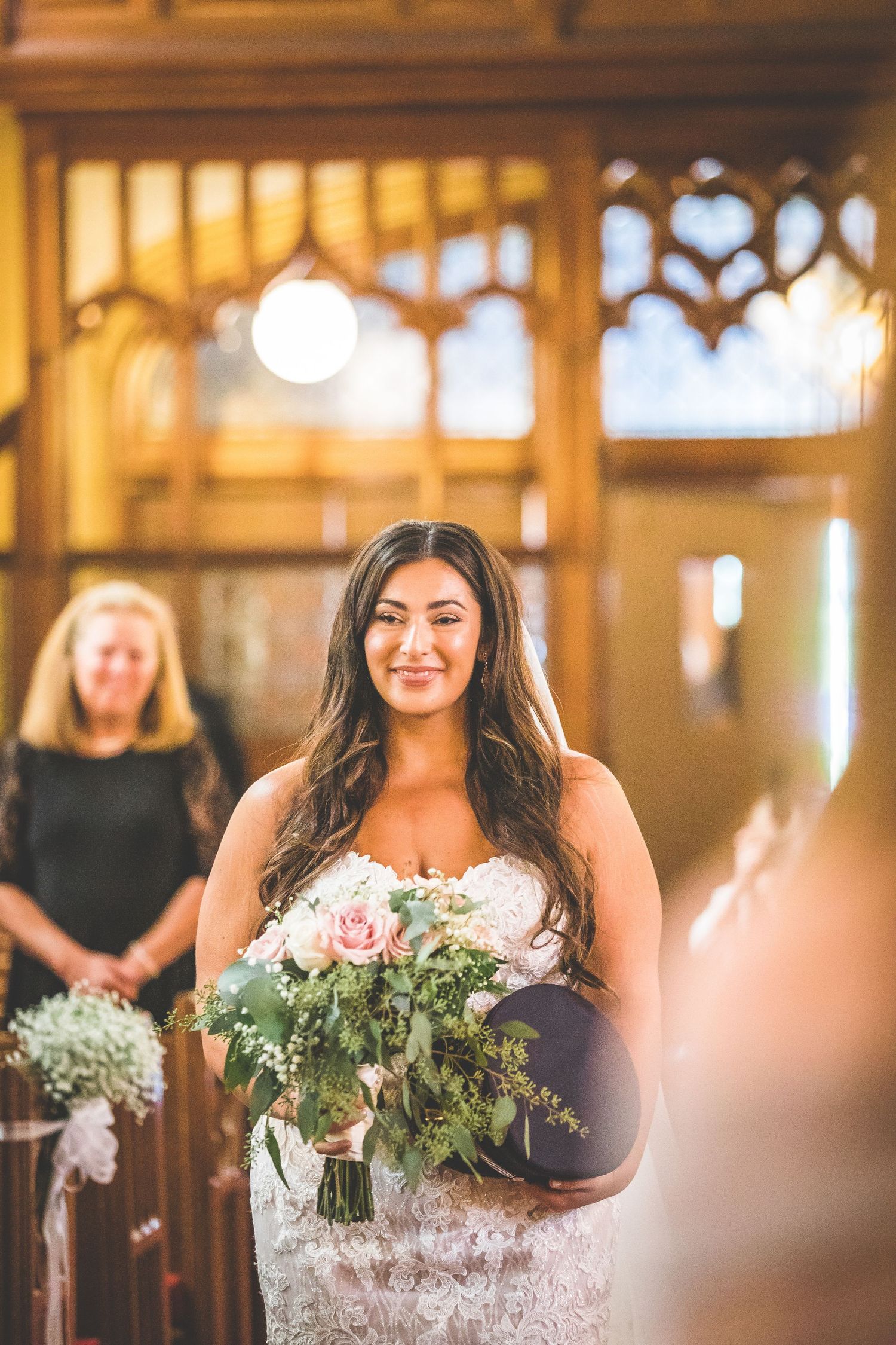 Bride holding floral bouquet stands in sunlit church aisle.