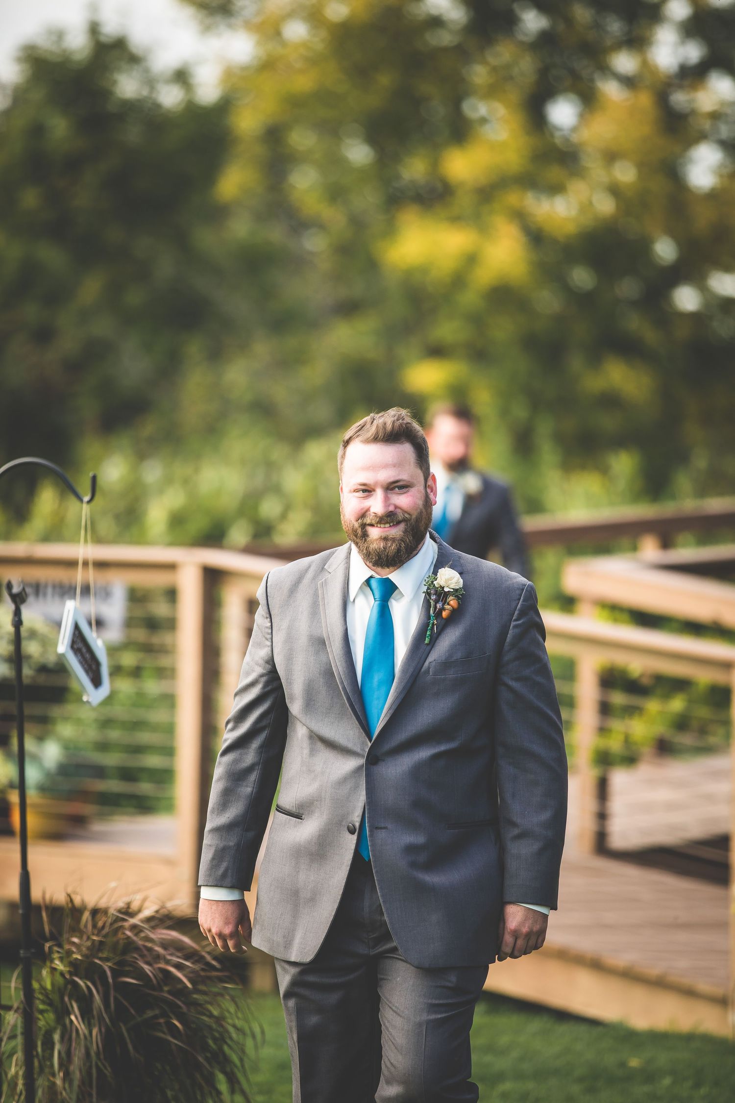 Groom in grey suit and bright blue tie walks along wooden bridge pathway.