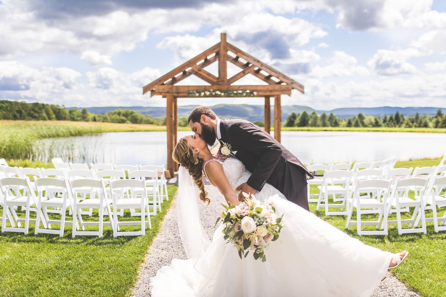 Wedding ceremony setup with wooden arch, white chairs, and couple sharing romantic moment.