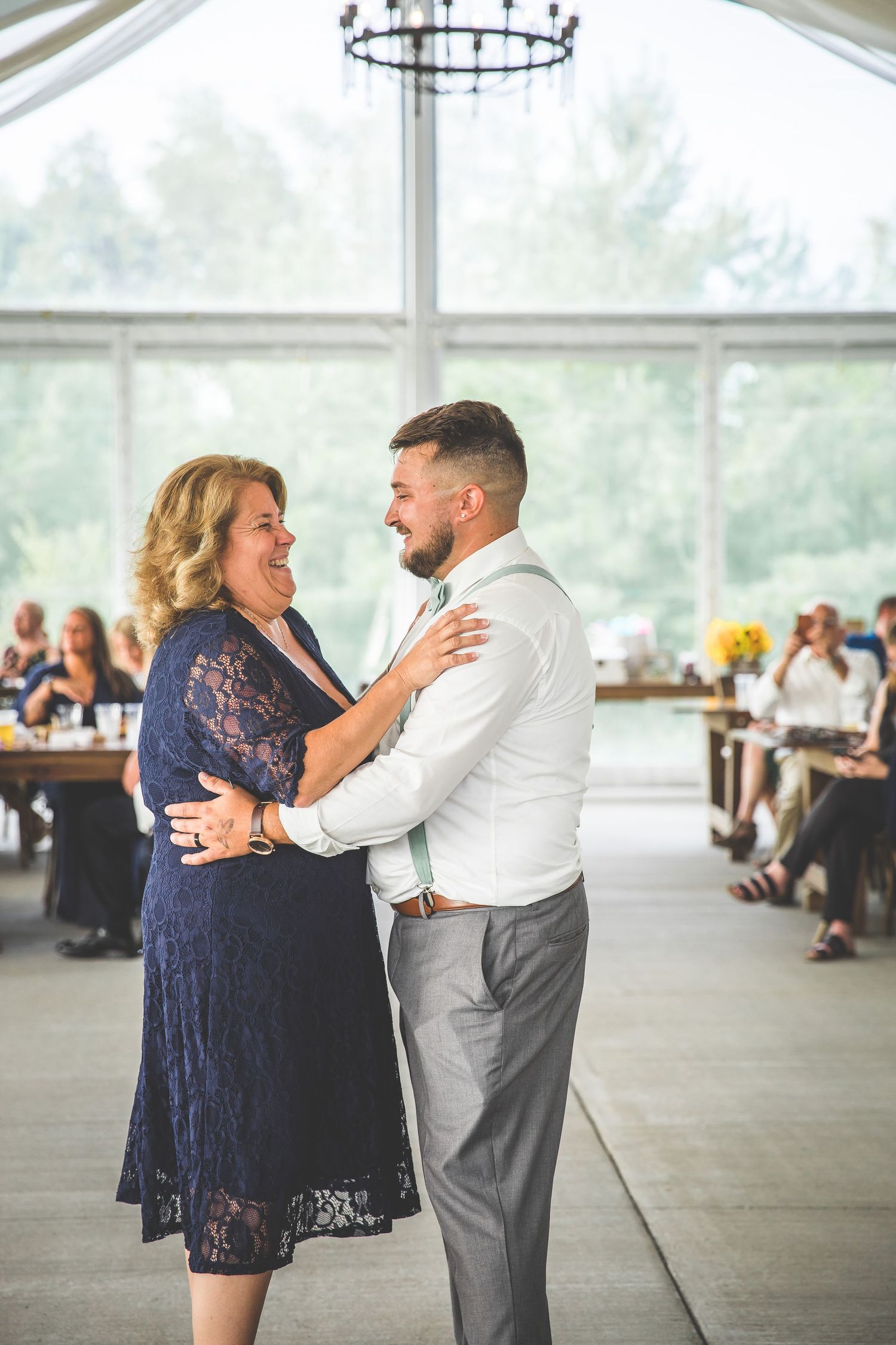 Two people share a joyful dance moment at a wedding reception in an elegant venue with large windows.