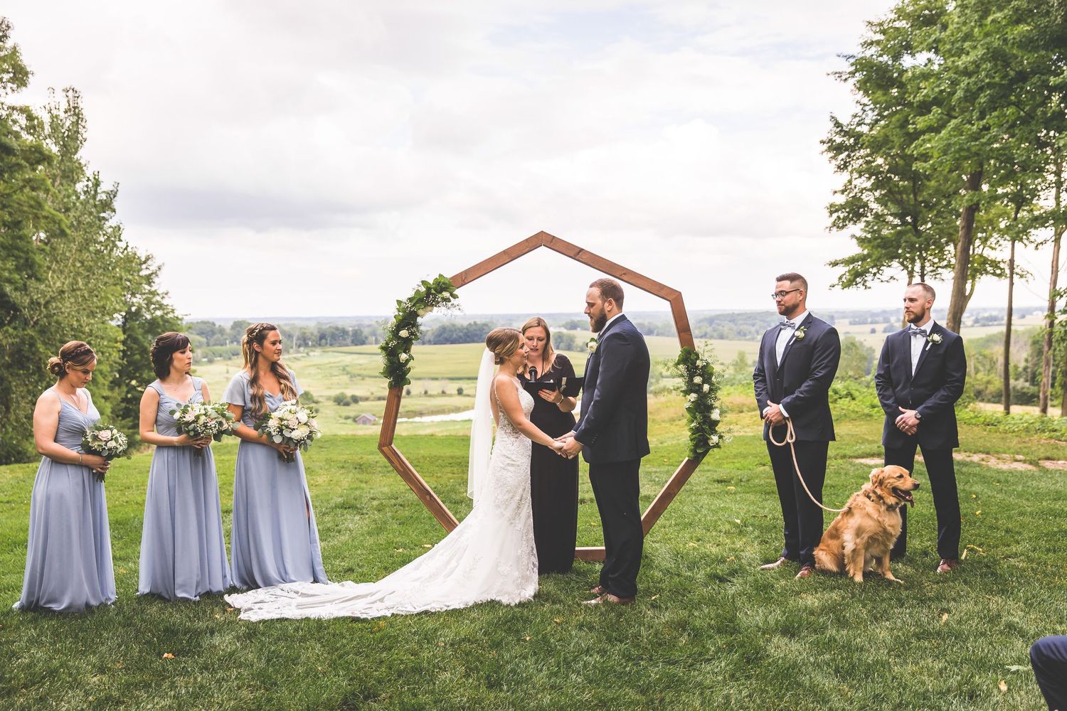Outdoor wedding ceremony under hexagonal arch with wedding party and golden retriever in attendance.