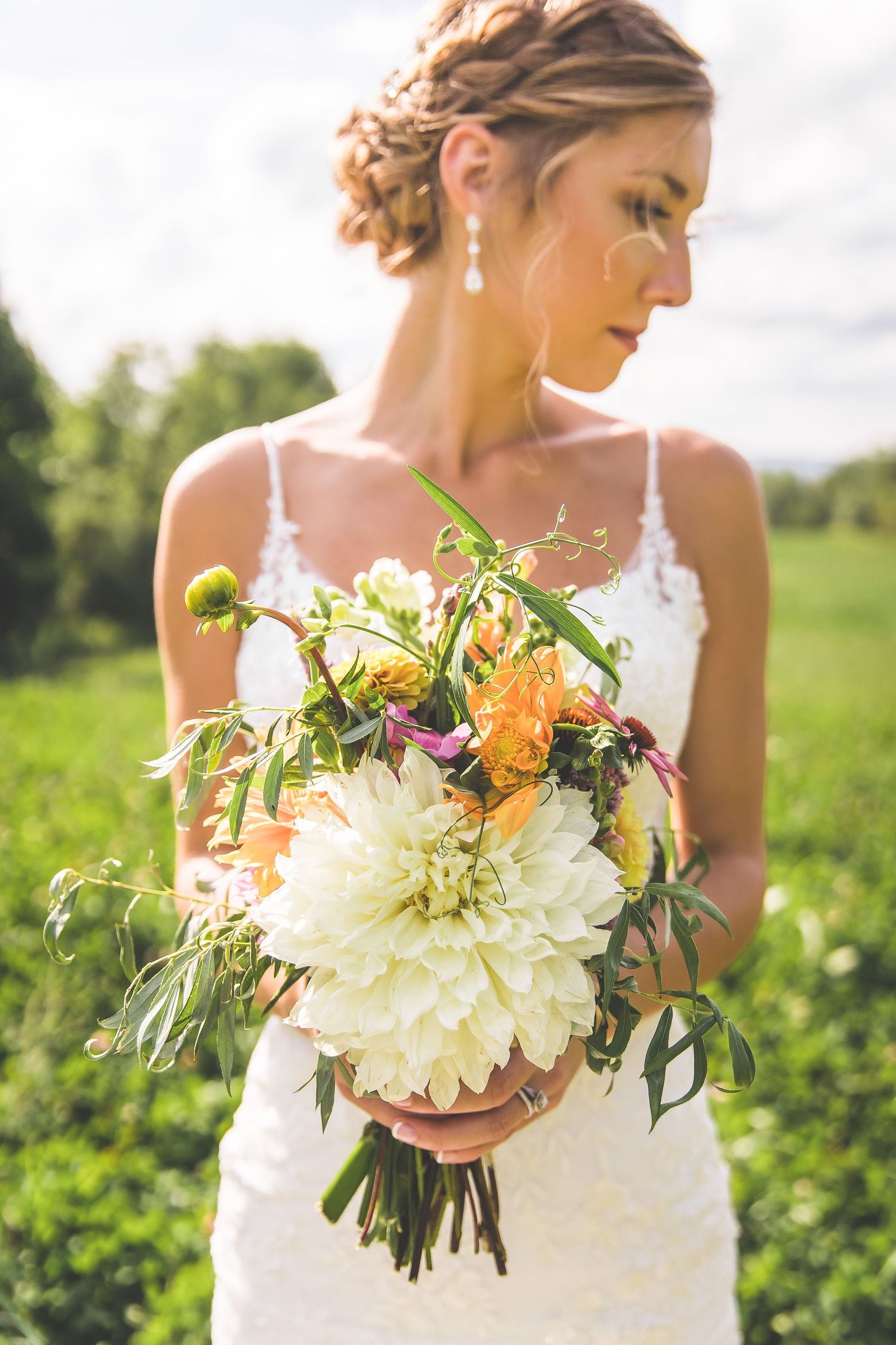 A wedding bouquet with white dahlias, orange flowers and greenery held against a white dress in a sunny field.