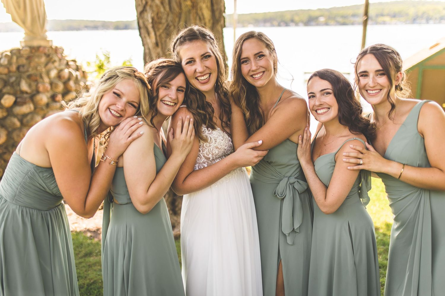Bridal party in sage green dresses shares intimate moment during lakeside wedding.