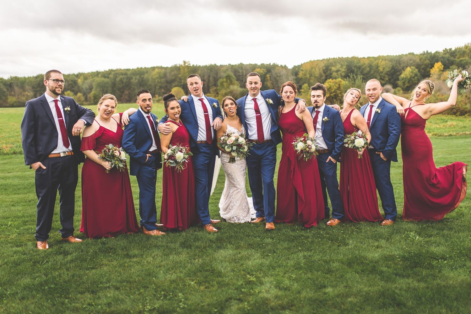 Large wedding party posing casually on grass with burgundy bridesmaids dresses and navy suits.