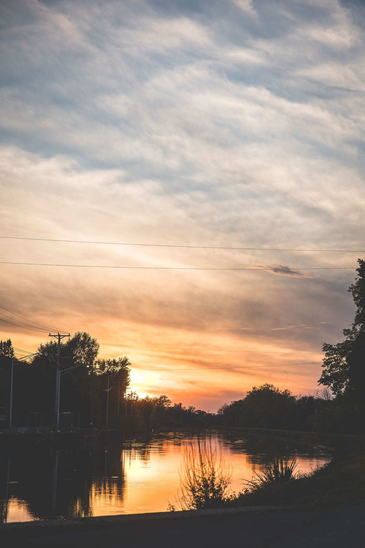 Scenic sunset over a lake with silhouetted trees and power lines against an orange and pink sky.