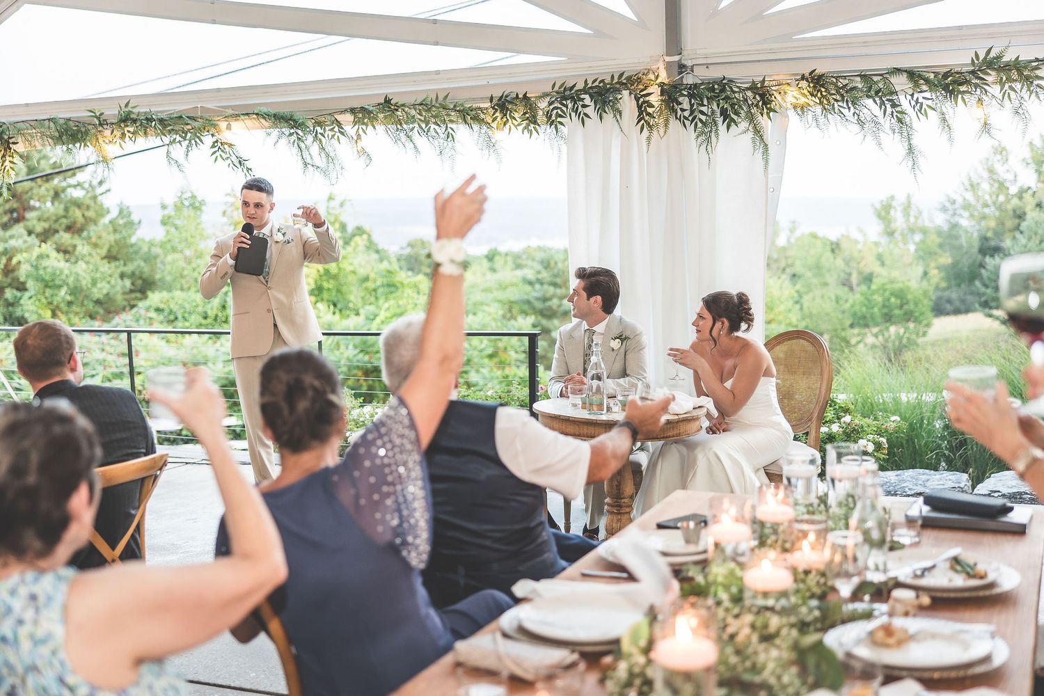 Wedding guests seated at tables raise their hands in celebration under a white tent.