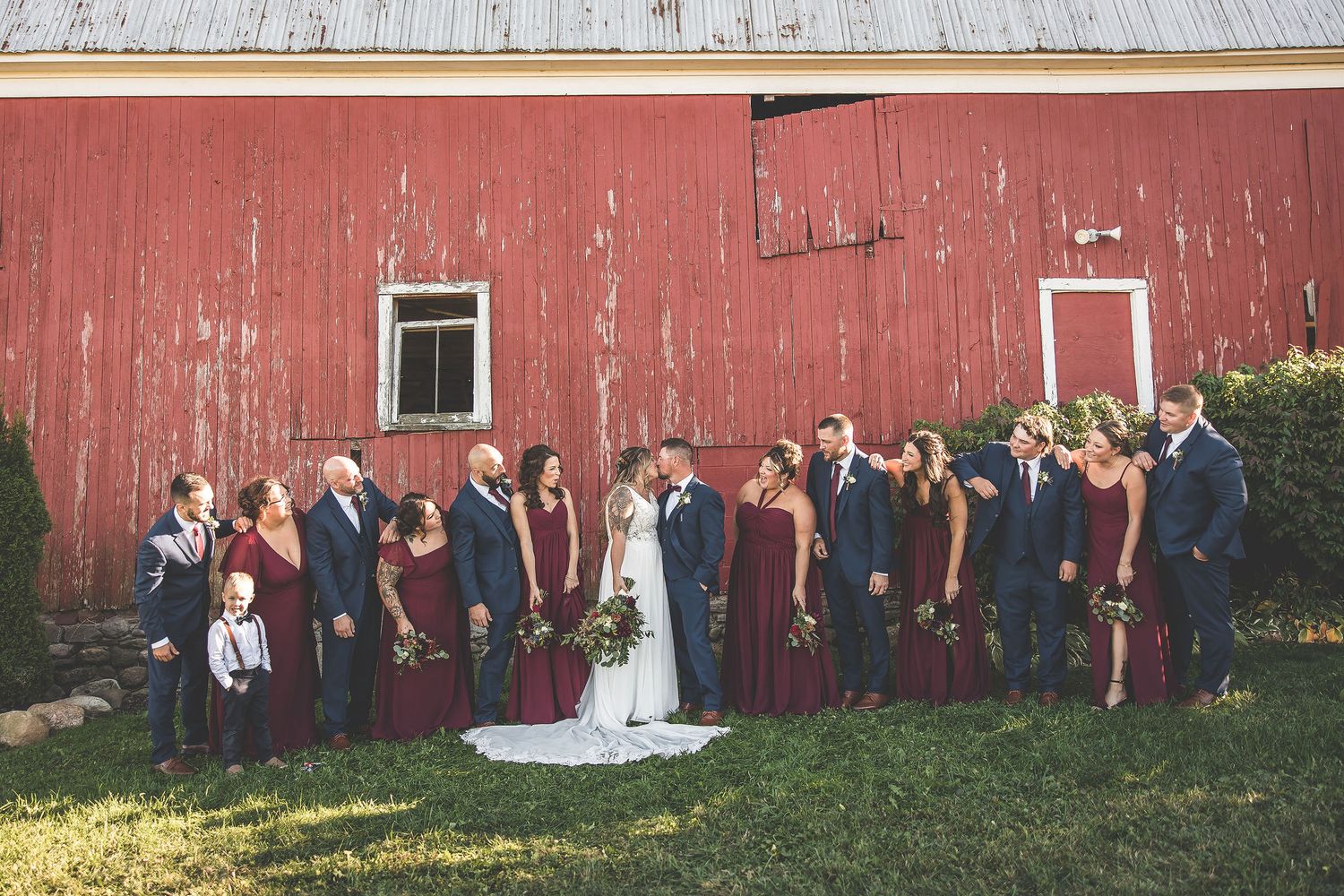 Full wedding party poses for wide-angle photo in front of rustic barn setting.