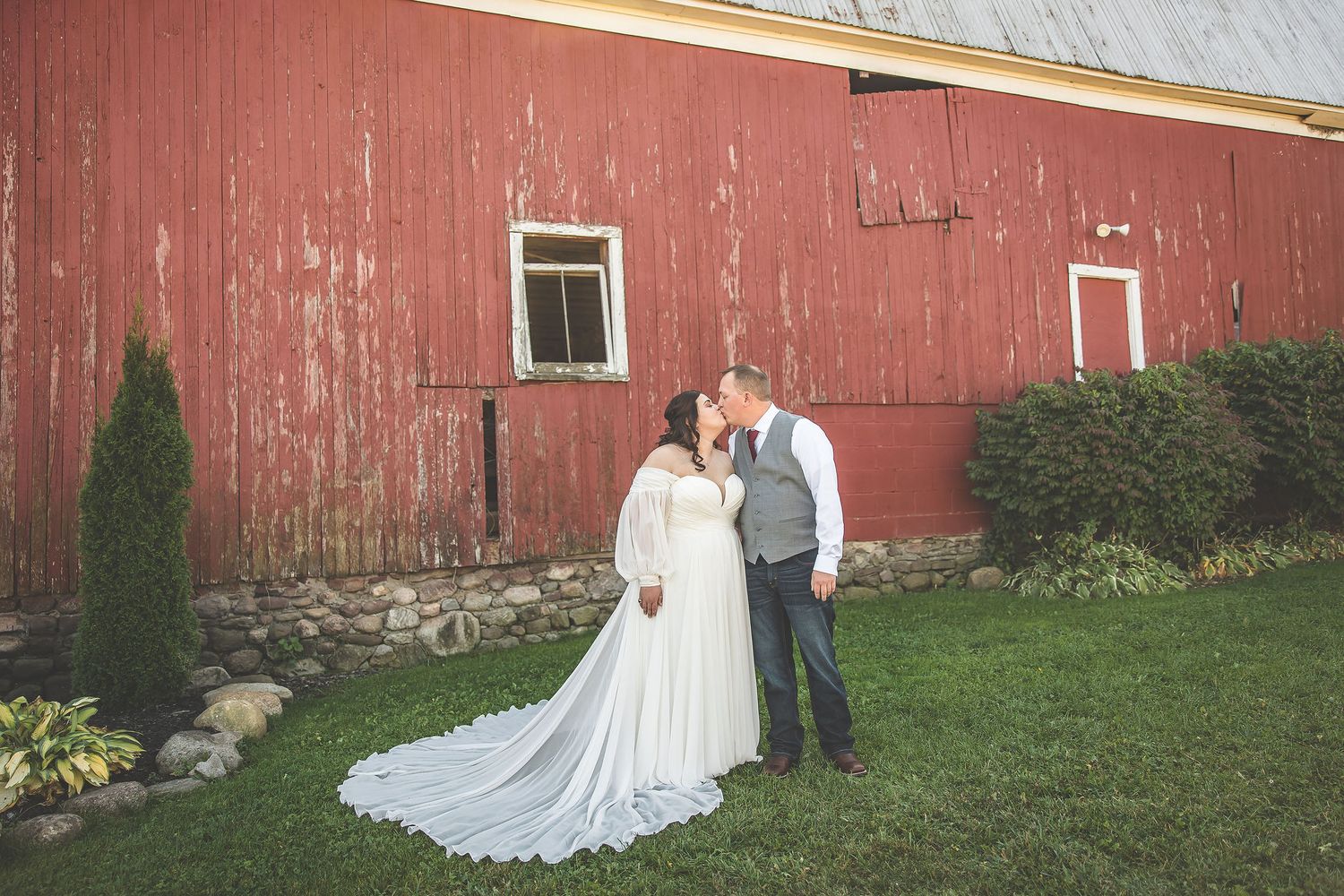 Wedding couple embraces beside rustic red barn in countryside setting.