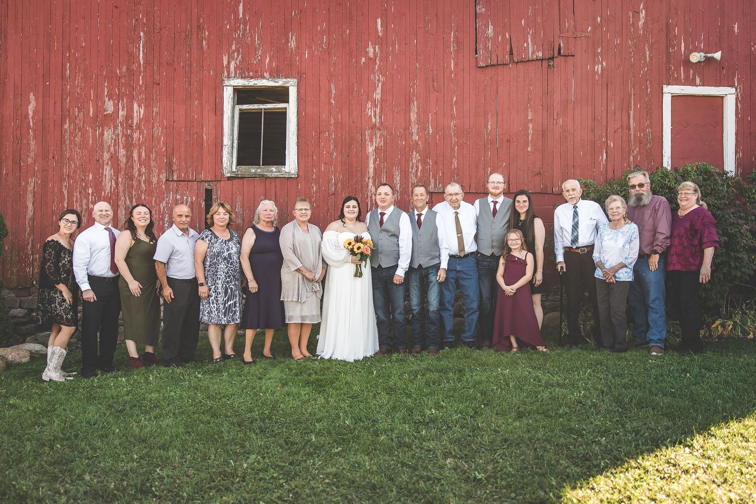 Wedding party and guests gathered for formal photos against weathered red barn siding.