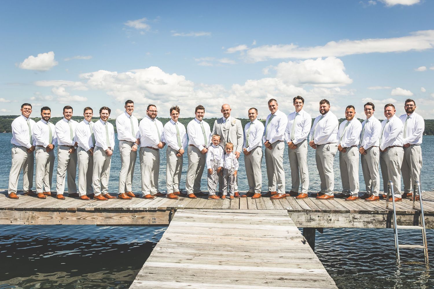 Wedding party in light gray suits and white shirts poses on wooden dock over water.