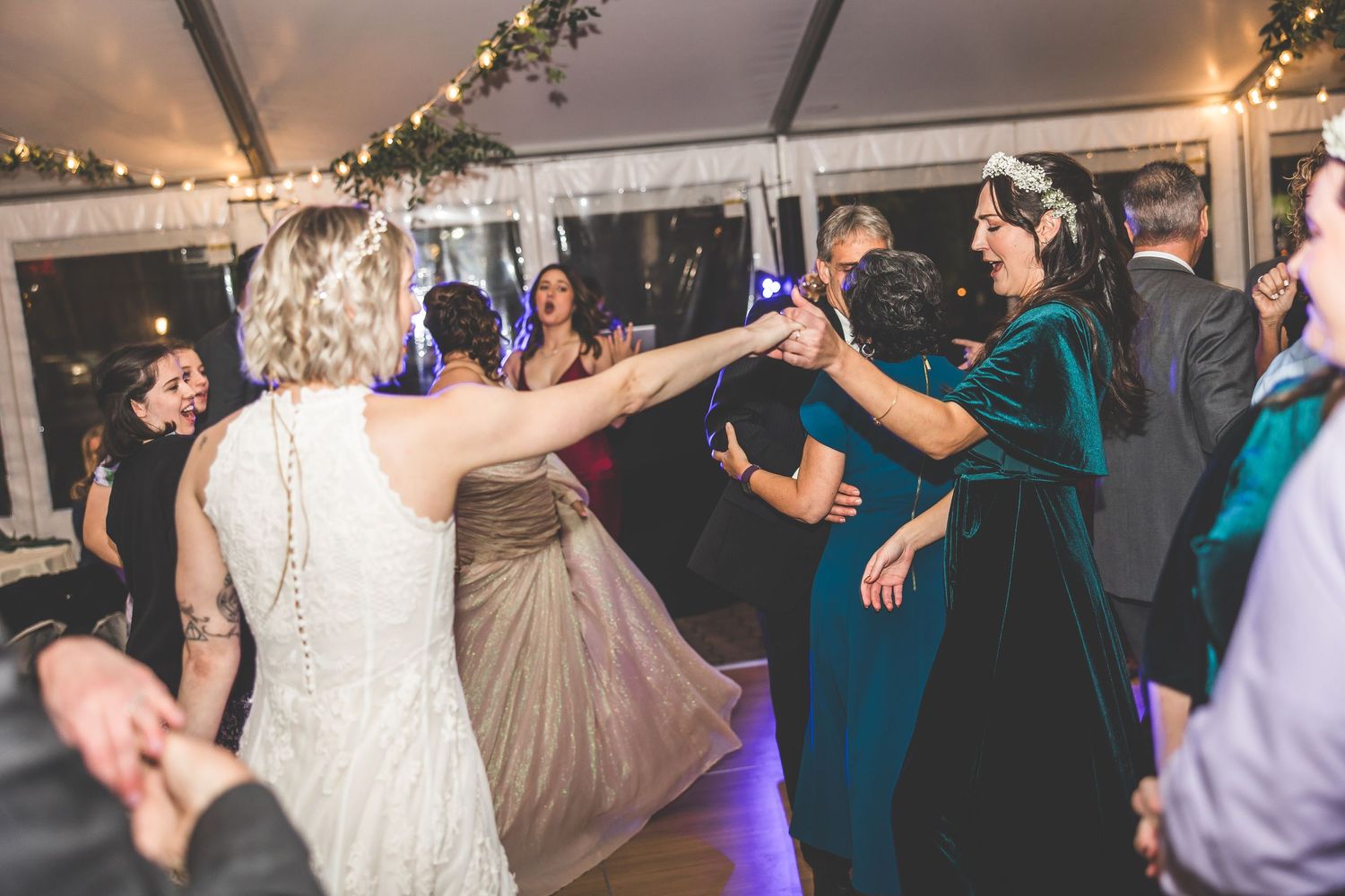 Wedding party and guests dance energetically at a tent reception with string lights.