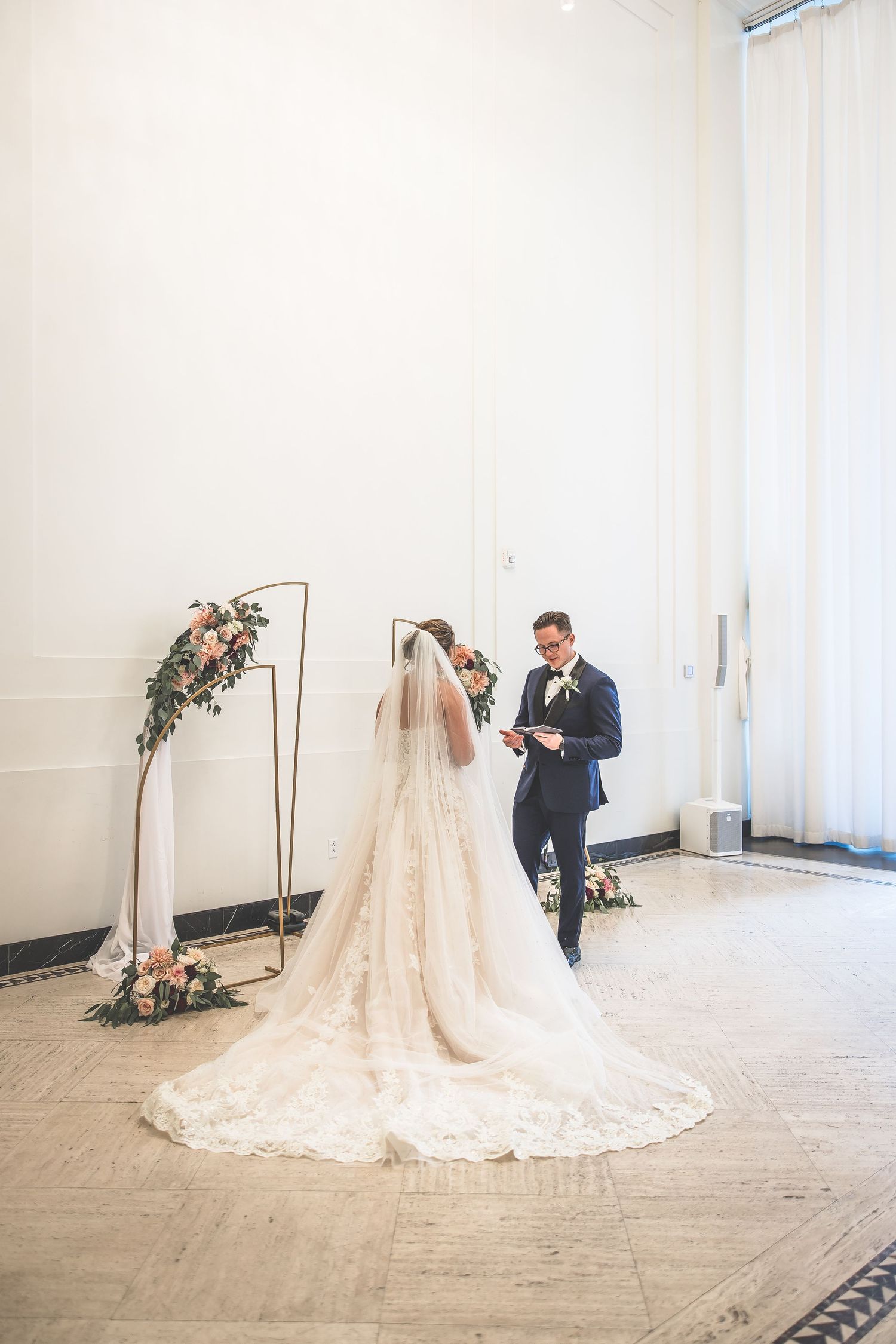 A bride in a lace wedding gown with long train stands before a minimalist floral arch during the ceremony.