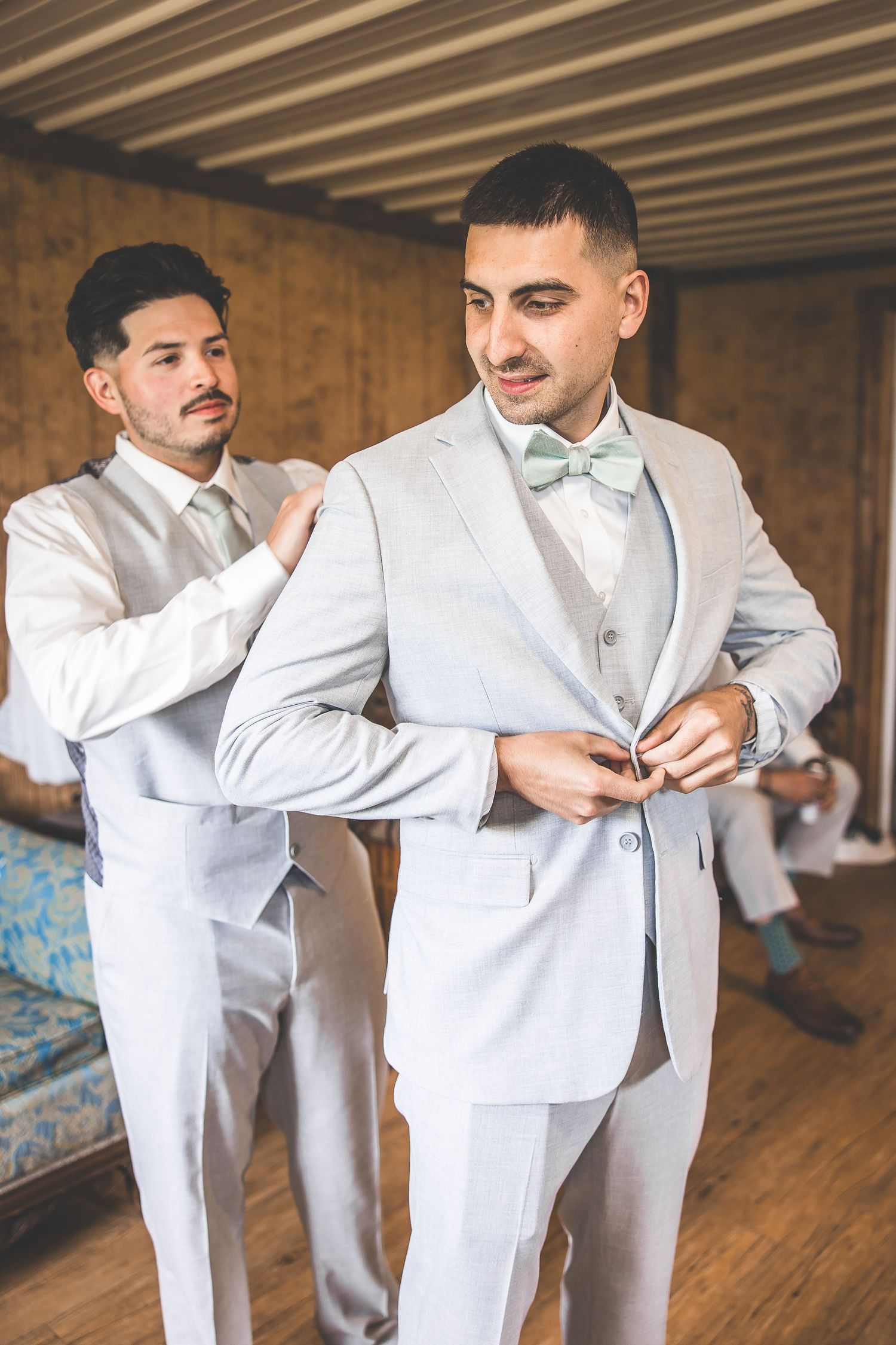 Groomsmen in light gray suits assisting each other with wedding day preparations.
