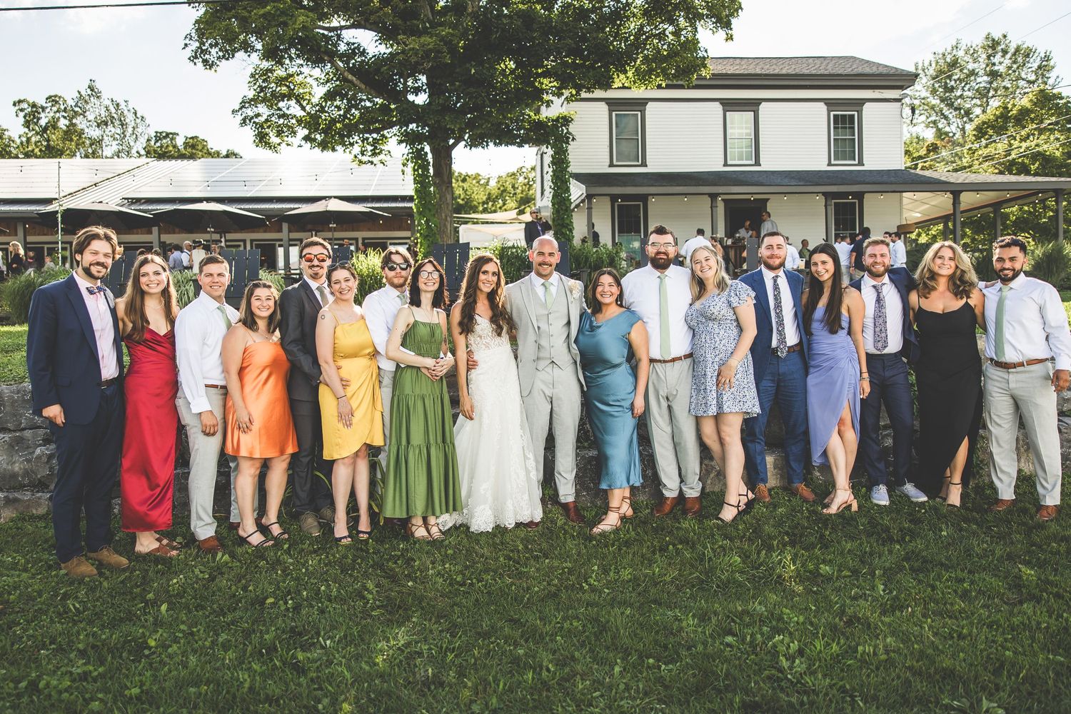 Wedding party and guests lined up for a large group photo on a grassy lawn.