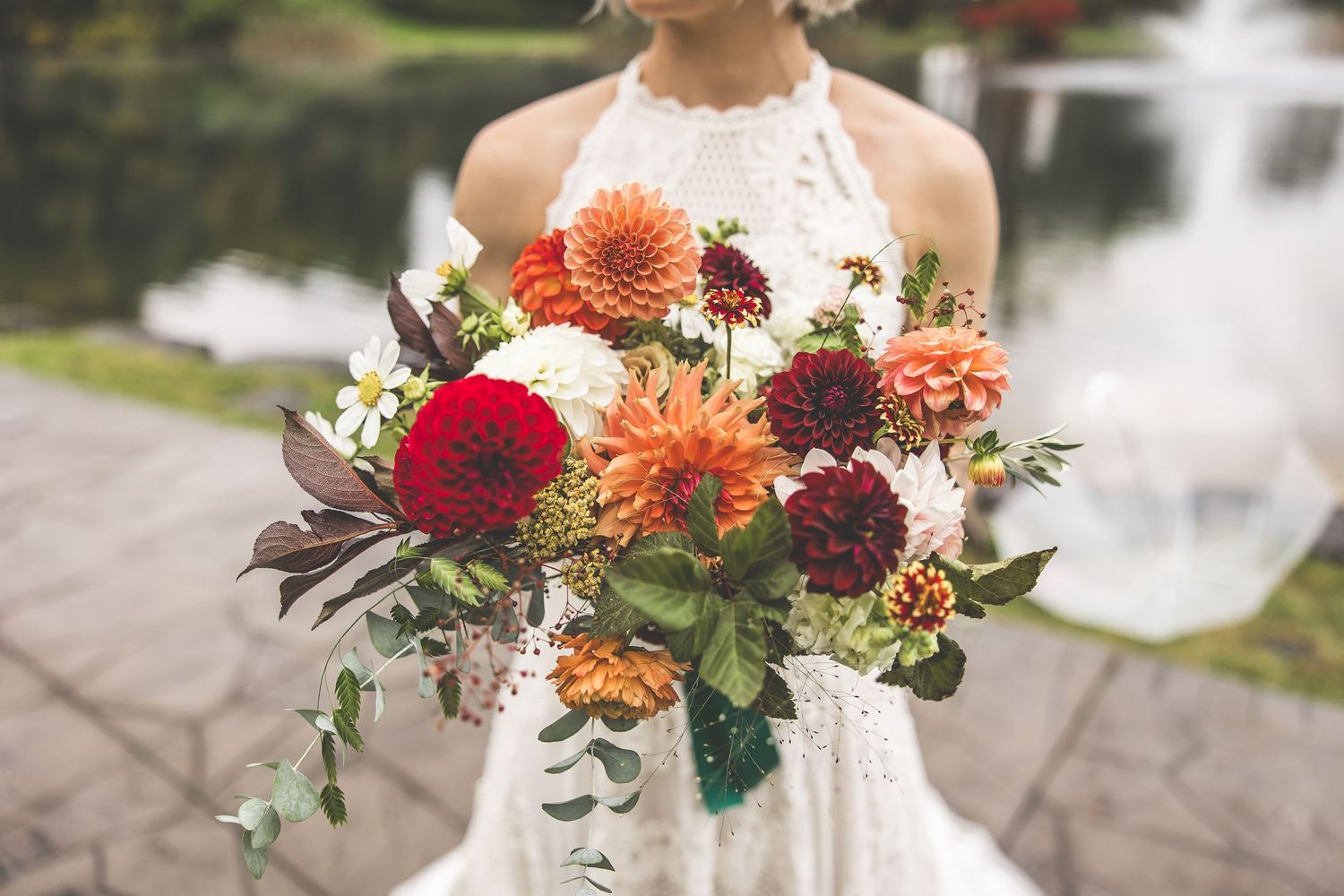 A colorful autumn wedding bouquet with dahlias, roses and greenery against a lake backdrop.