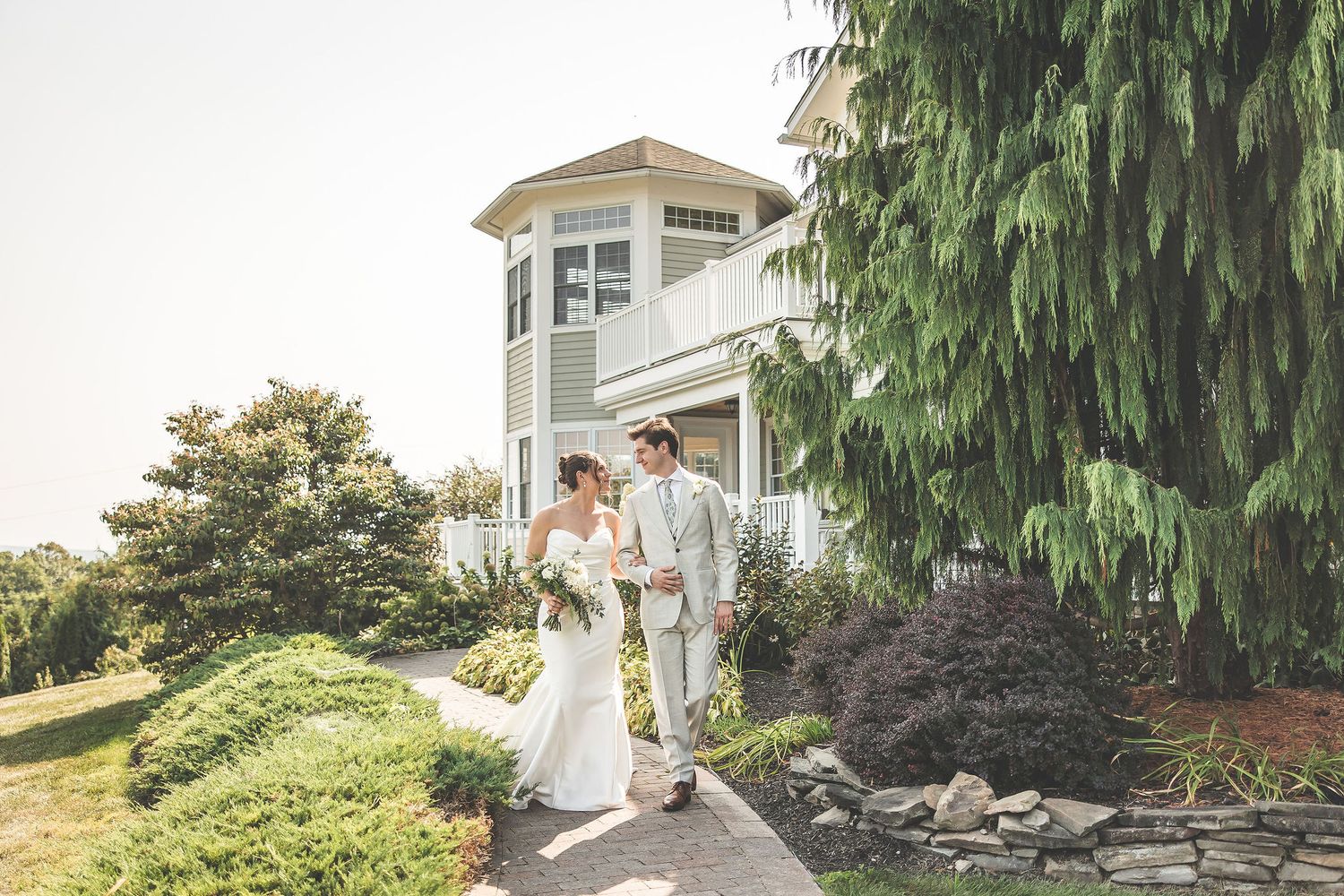 Couple poses near a grand coastal house surrounded by lush landscaping.