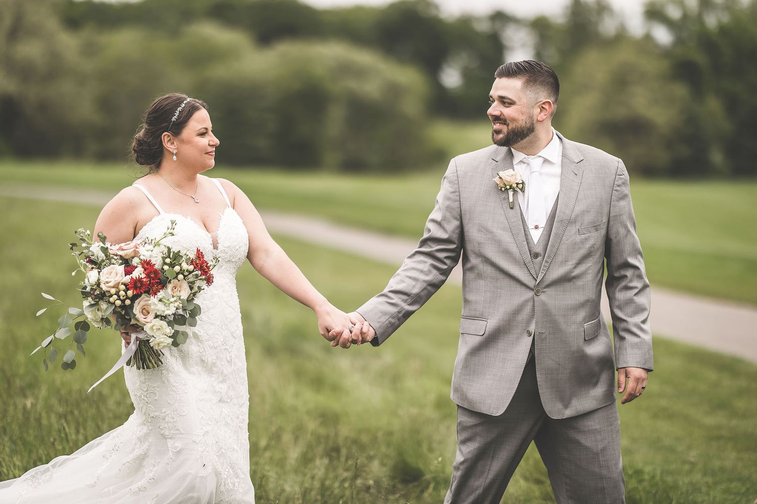 A joyful wedding couple holding hands while walking along a winding path through a lush green field.