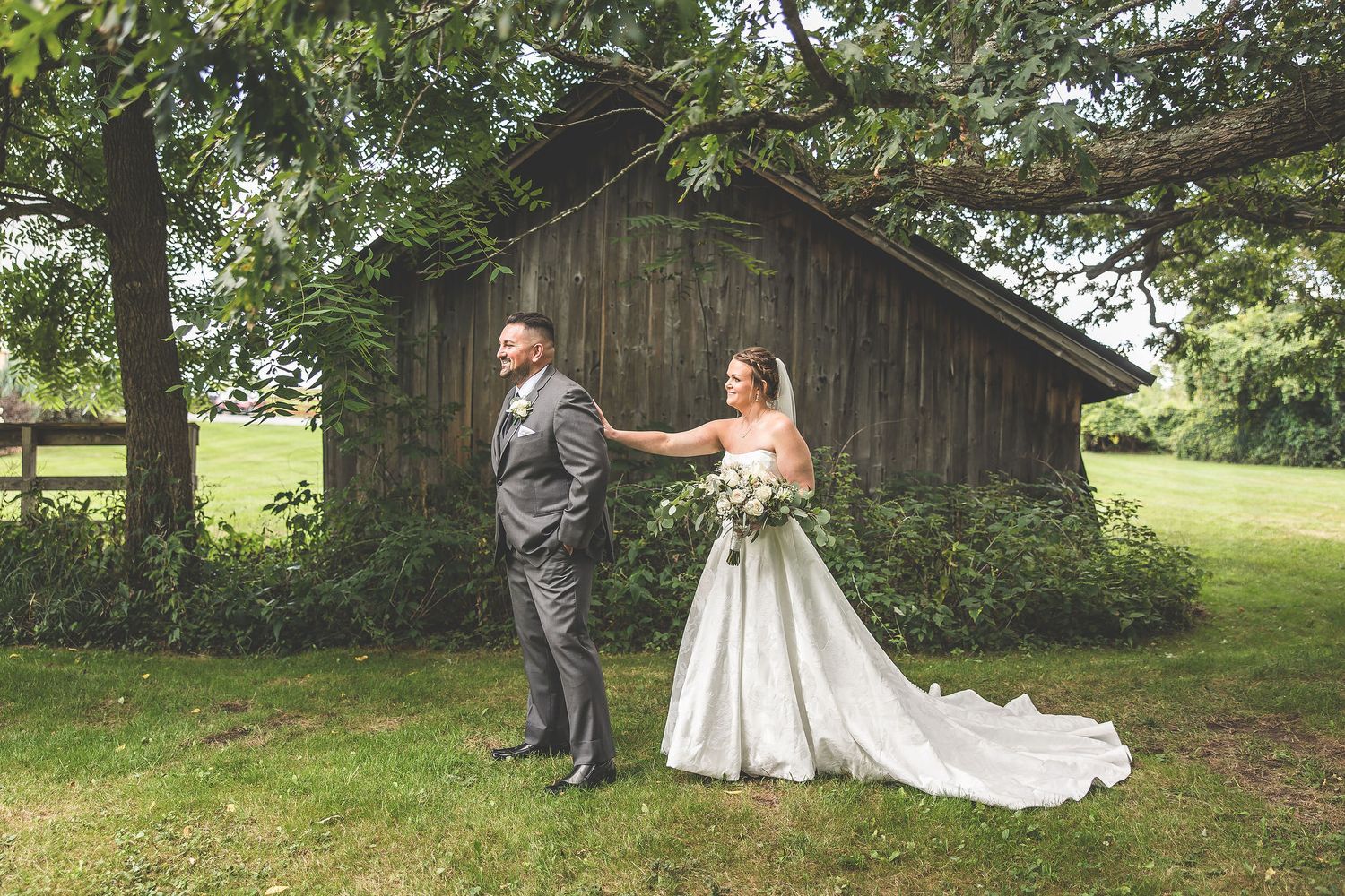 Wedding couple and bridal party pose in front of rustic barn on grassy hillside.