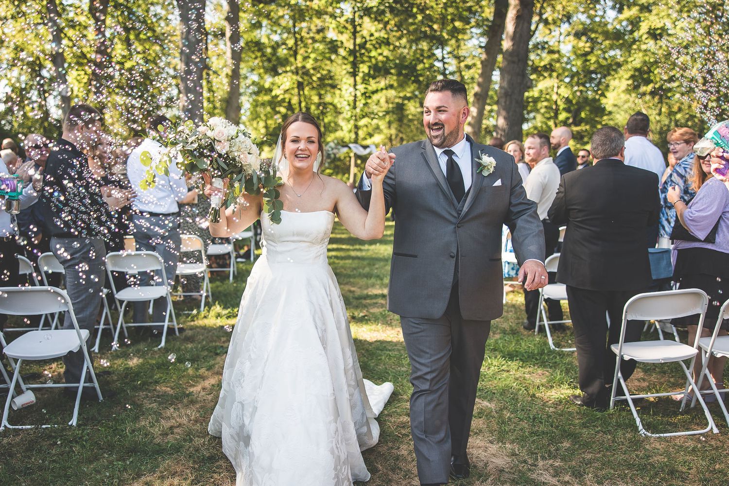 Outdoor wedding celebration in progress with guests seated among trees on a sunny afternoon.
