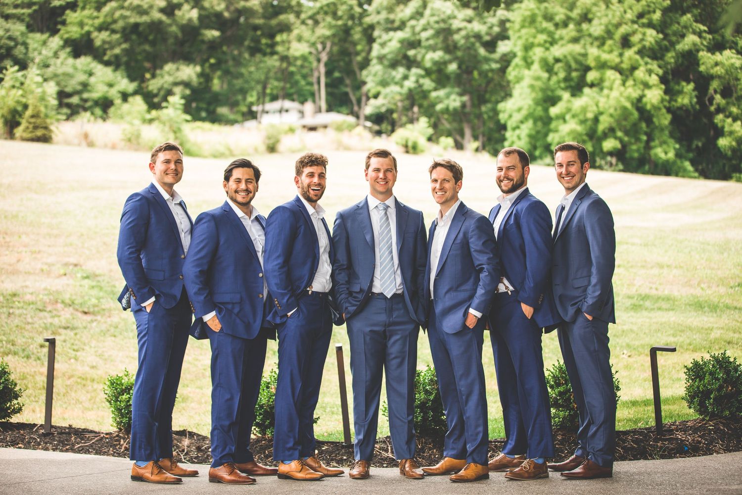 Wedding party in navy blue suits and dresses pose together outdoors on a sunny day.