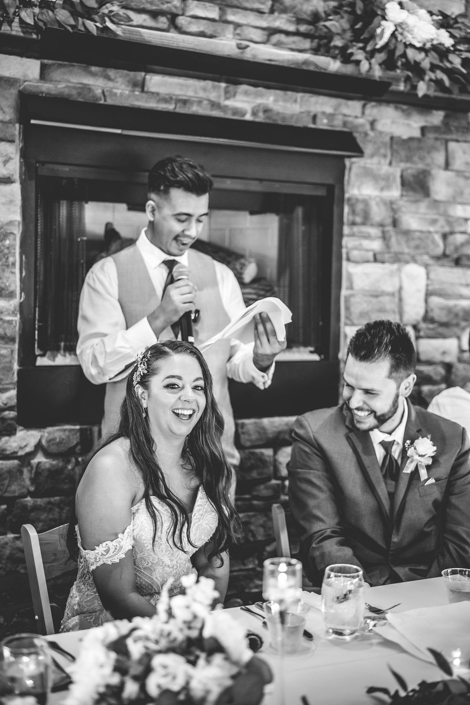 Candid black and white photo of wedding party enjoying speeches at reception.