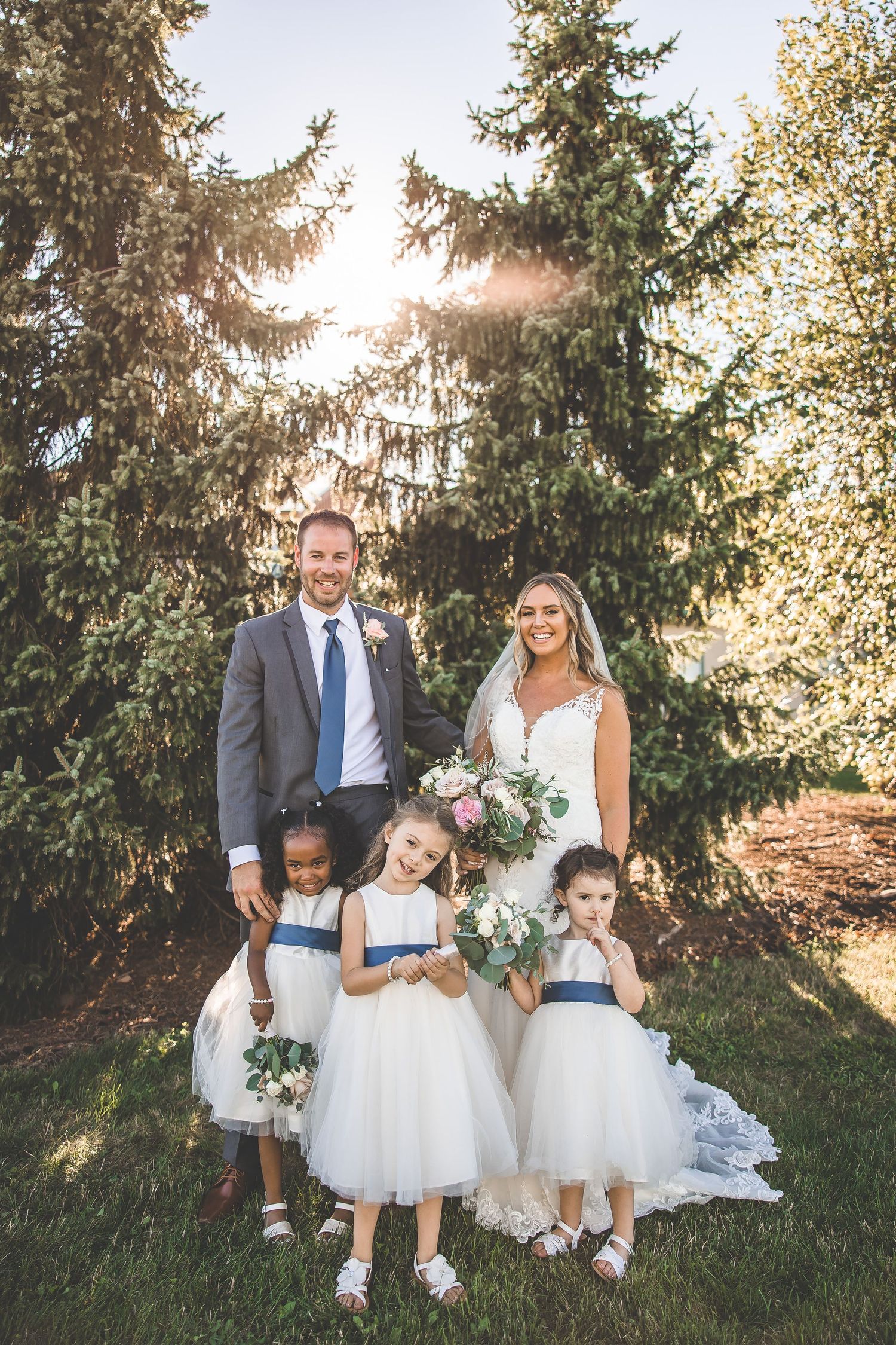 Wedding party portrait with flower girls in white dresses standing in front of tall evergreen trees.
