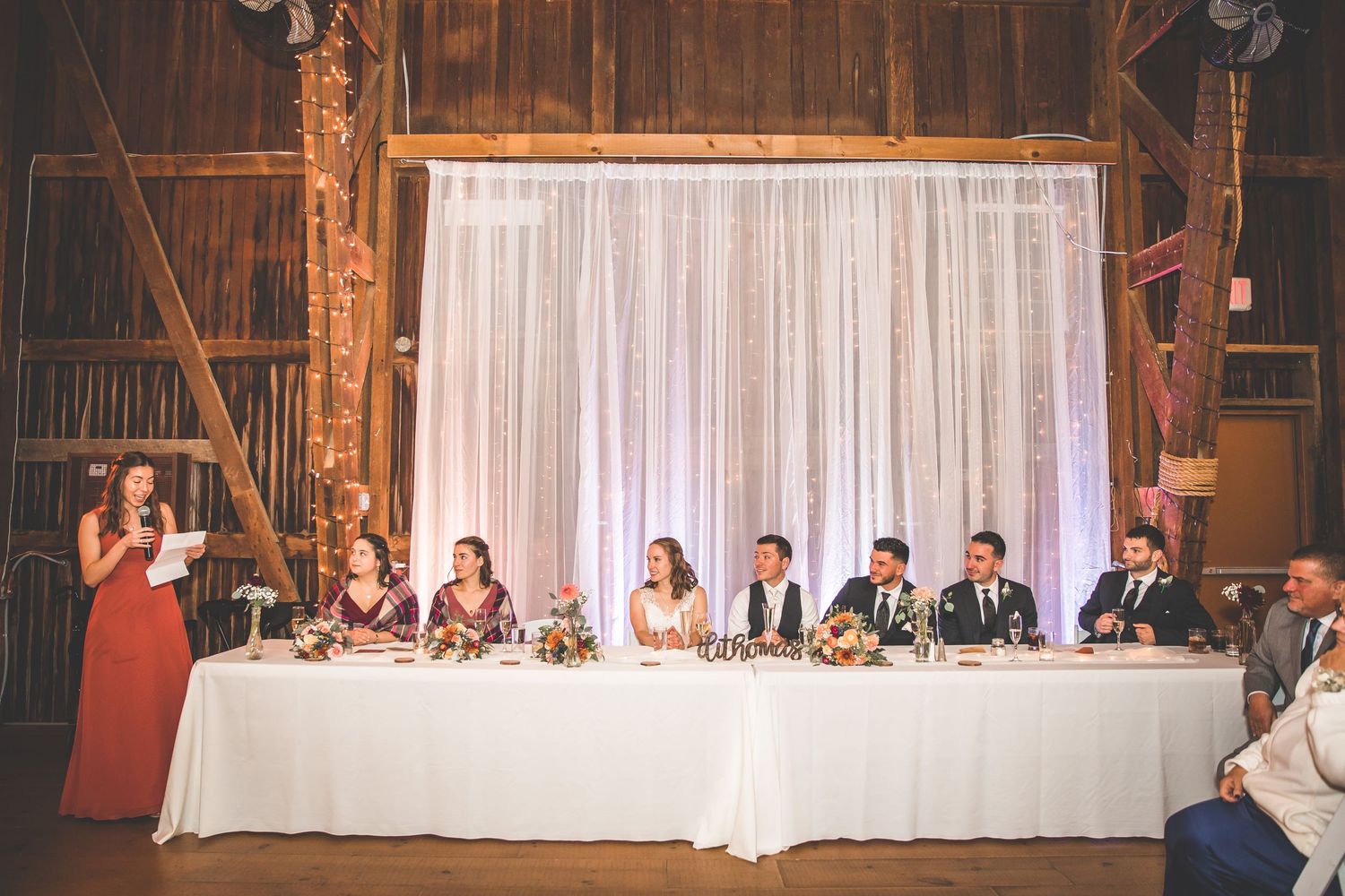 Long head table setup in a barn venue with white draping and warm lighting.