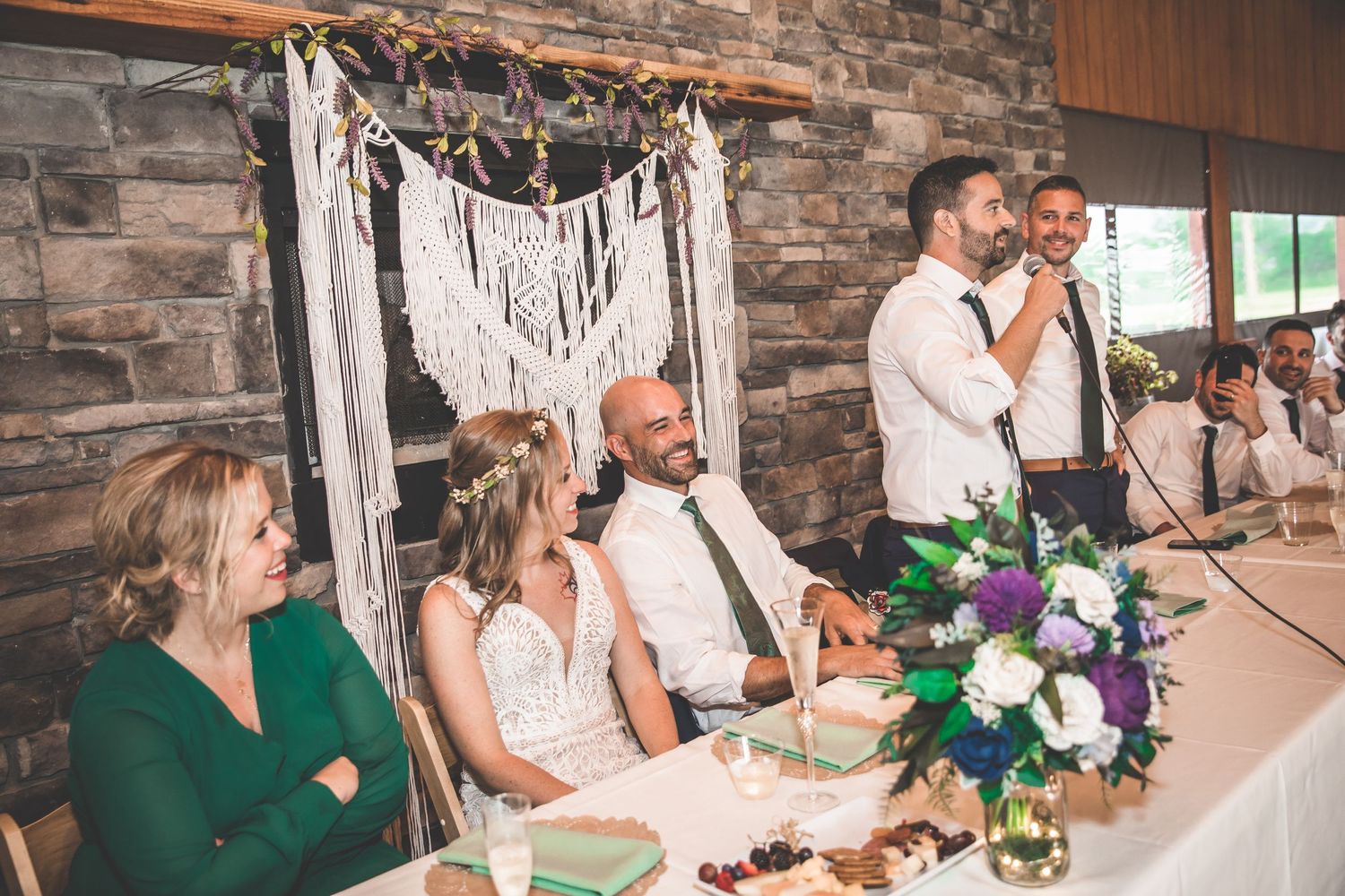 Wedding reception scene with stone walls and macrame backdrop during speeches.