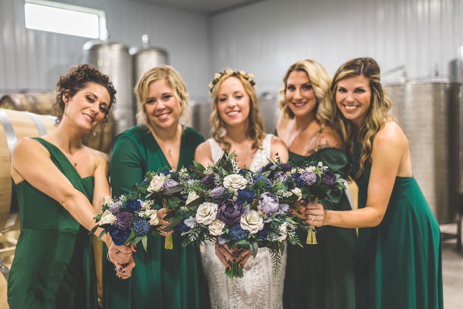 Bridesmaids in emerald green dresses pose together with bouquets in a winery setting.