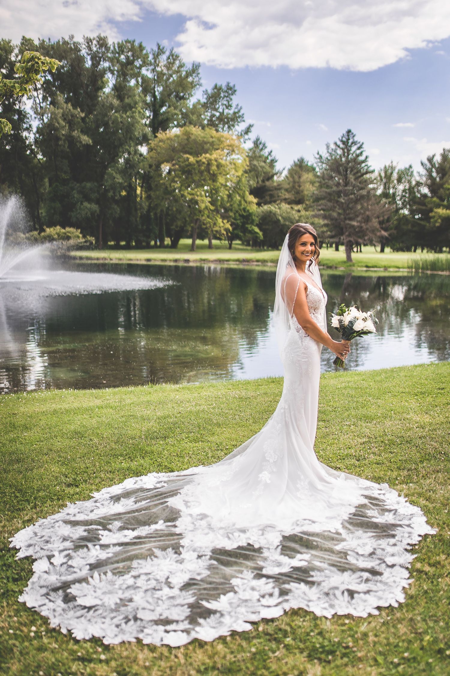 Wedding dress with long lace train photographed by a lake with fountain.