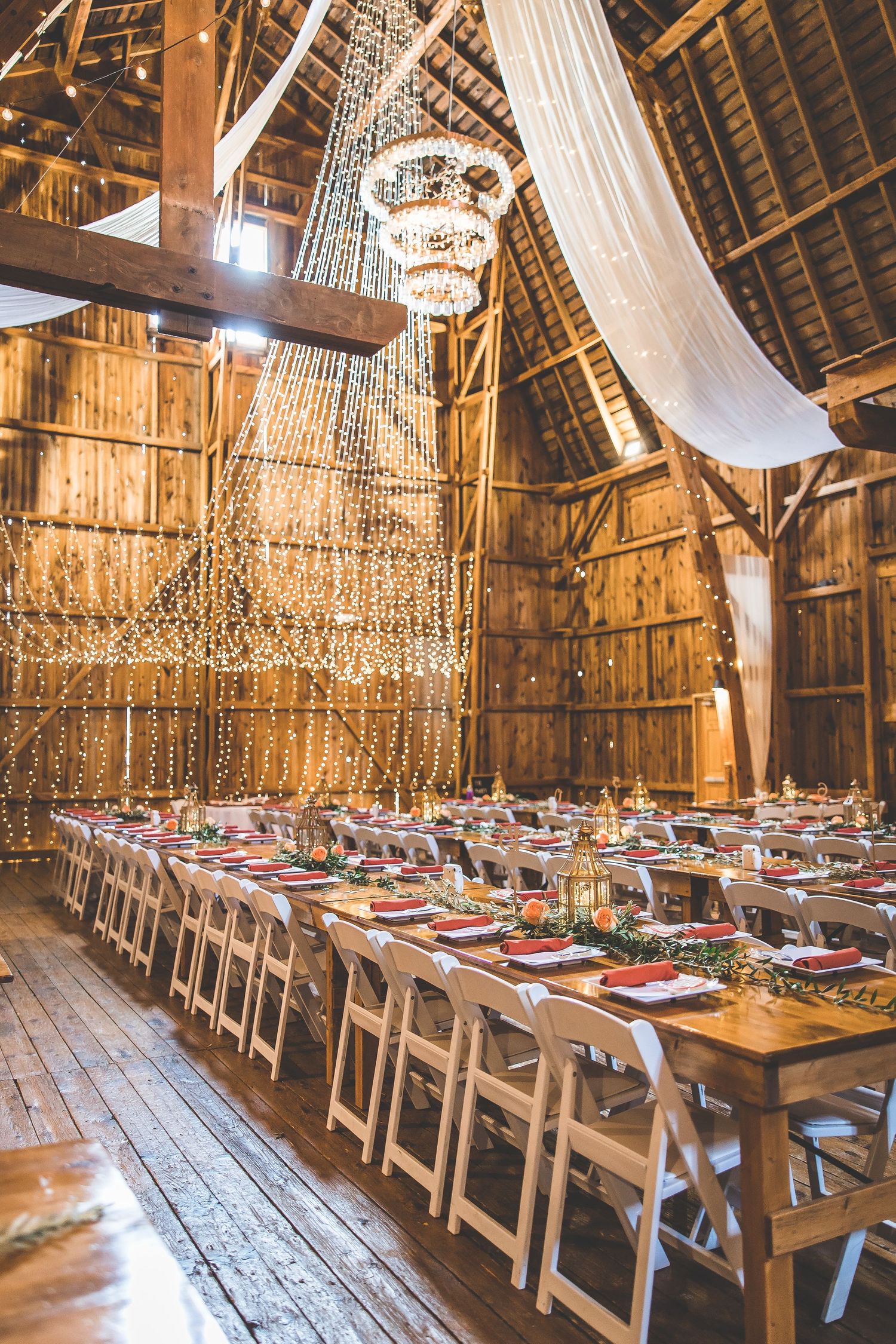 Rustic barn wedding reception setup with long tables and crystal chandeliers hanging from wooden beams.