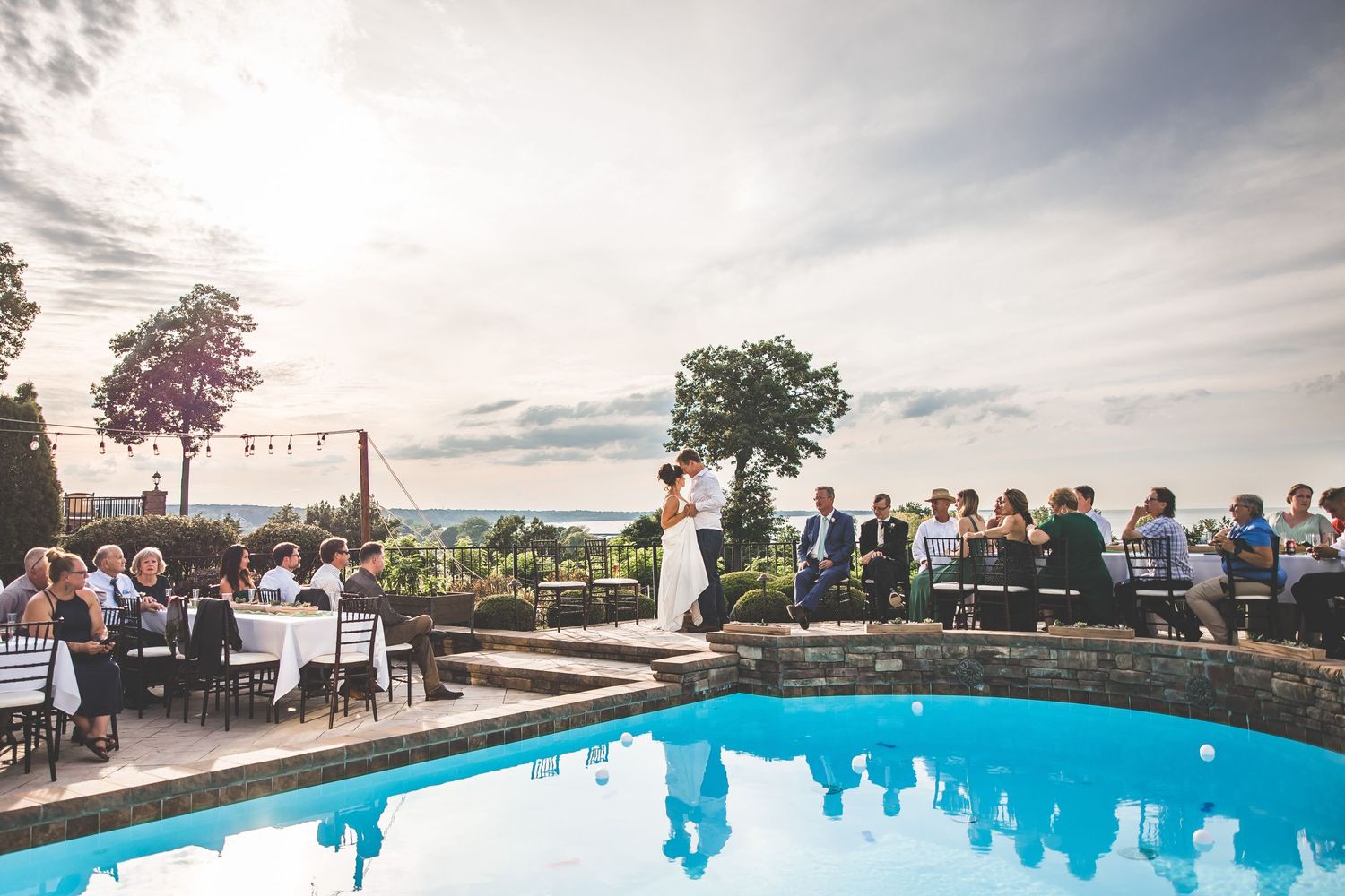Wedding guests gather around a swimming pool during an outdoor reception at sunset.