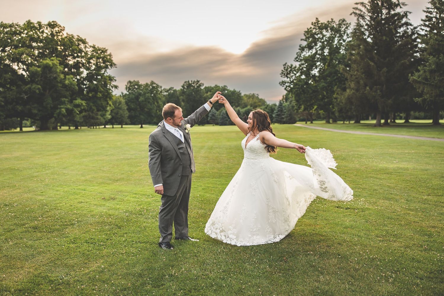 Wedding couple dances and twirls on a green lawn during golden hour.
