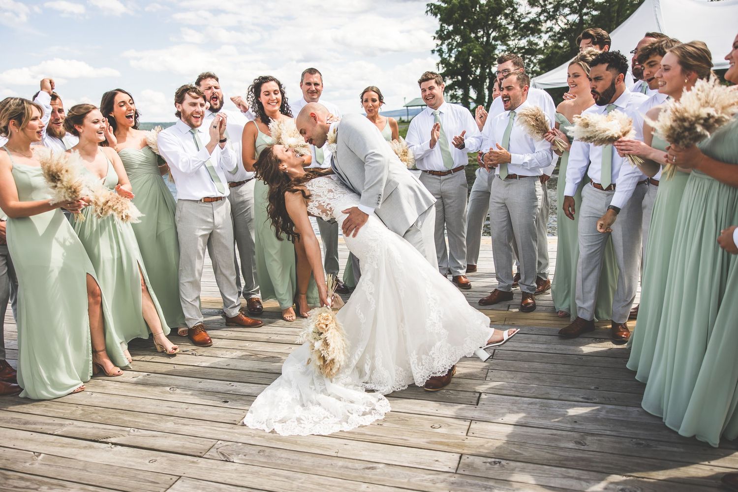 Wedding couple shares romantic dance moment surrounded by wedding party on wooden dock.