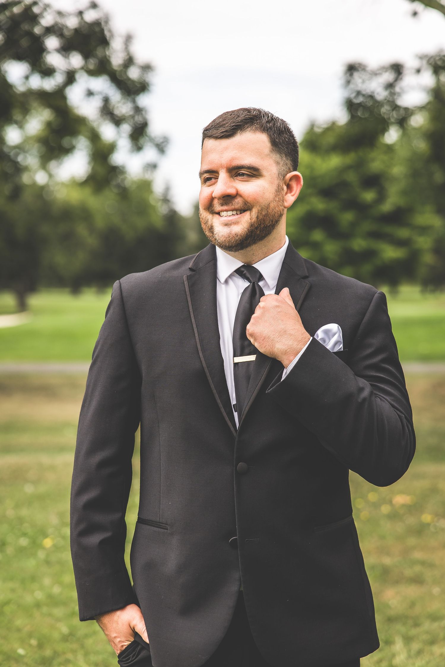 Groom in black tuxedo poses outdoors in a grassy field with natural lighting.