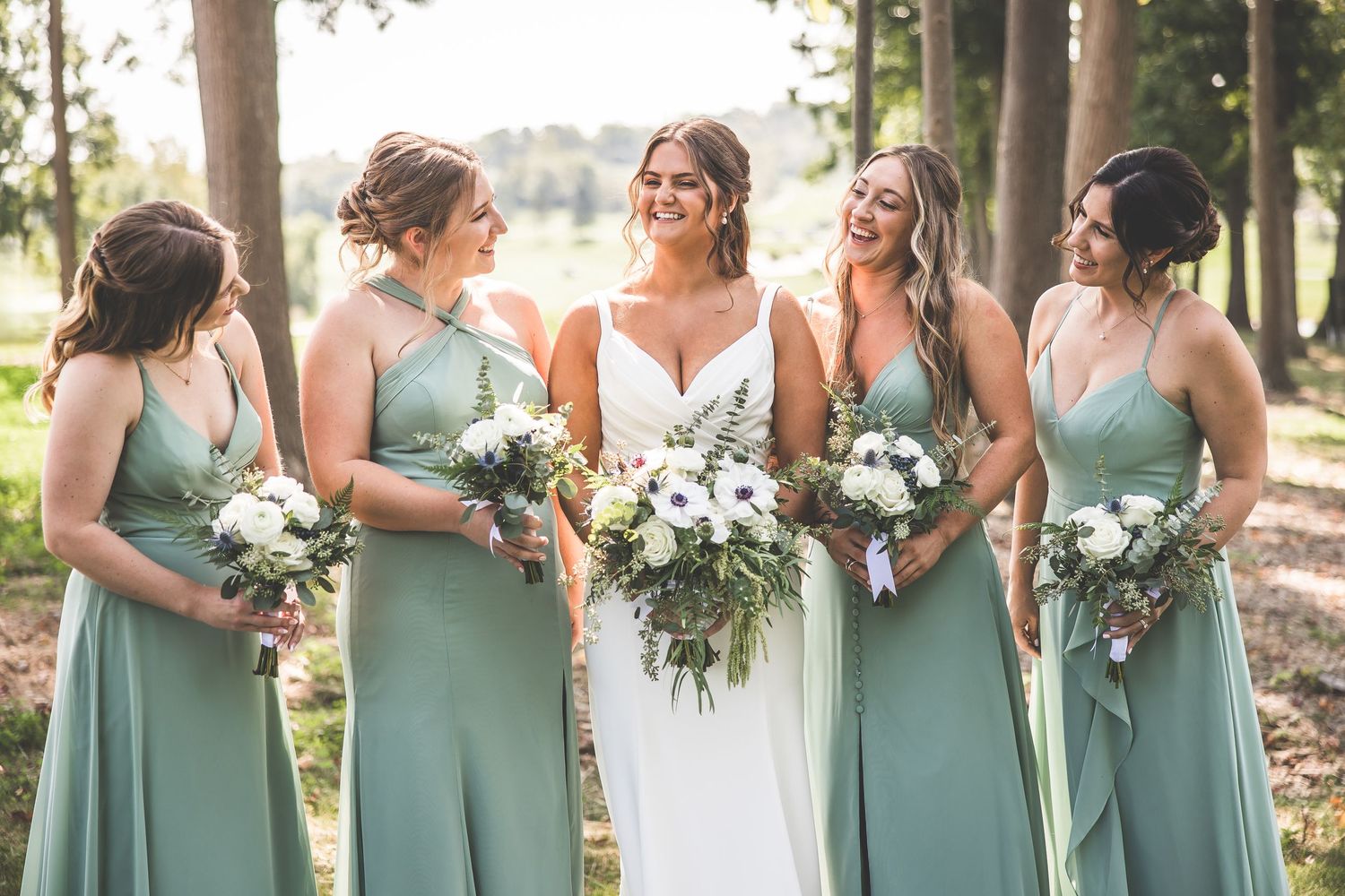 Bridal party in matching sage dresses stands with bouquets in an outdoor woodland setting.