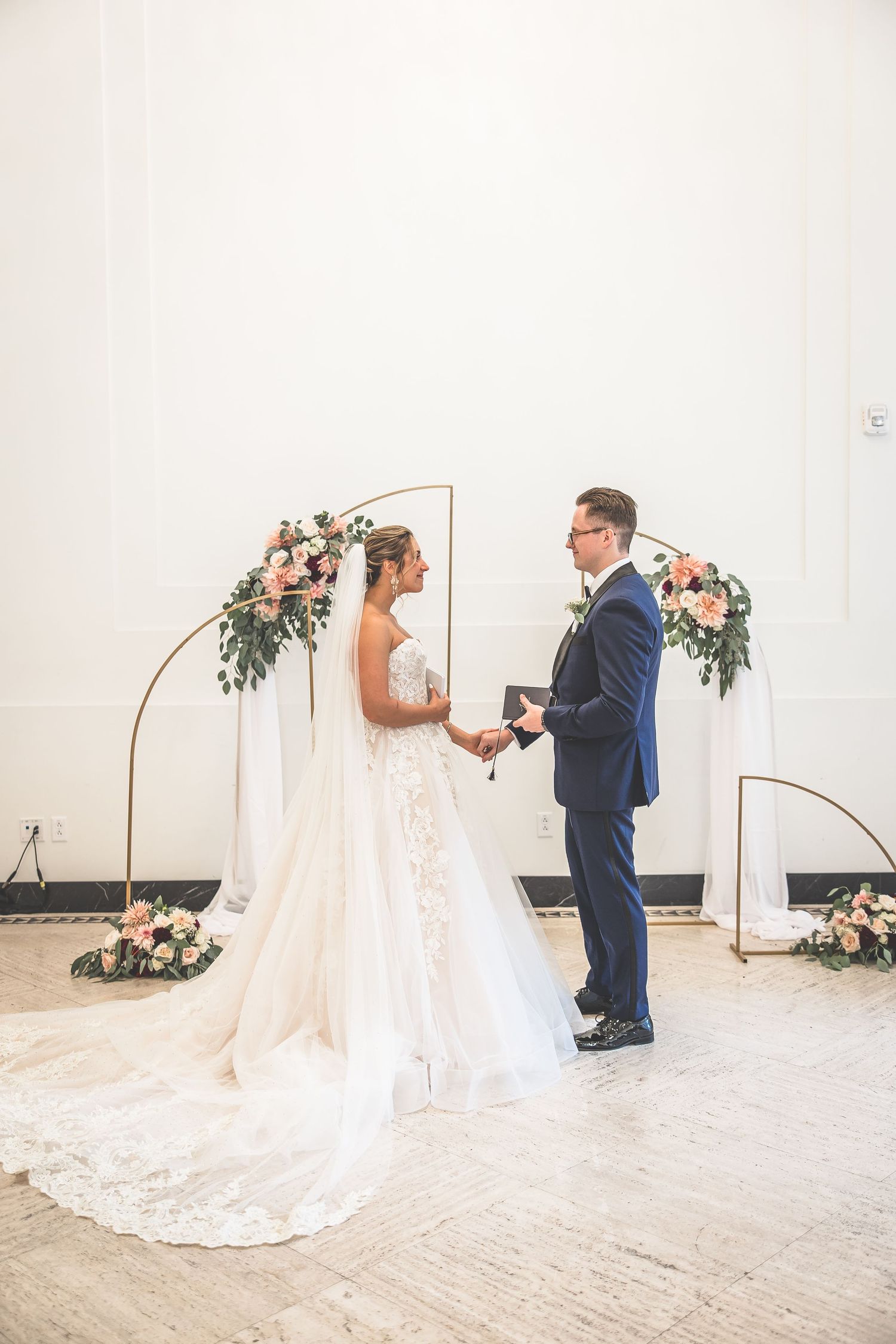 A couple stands beneath a geometric arch adorned with pink florals during their wedding ceremony.