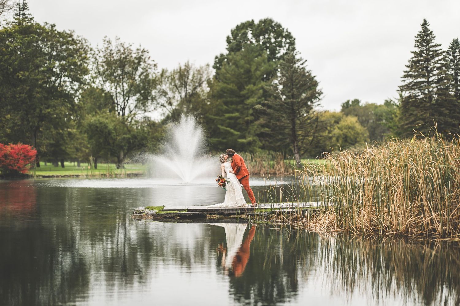 Couple poses on dock with water fountain creating beautiful reflection in serene lake setting.