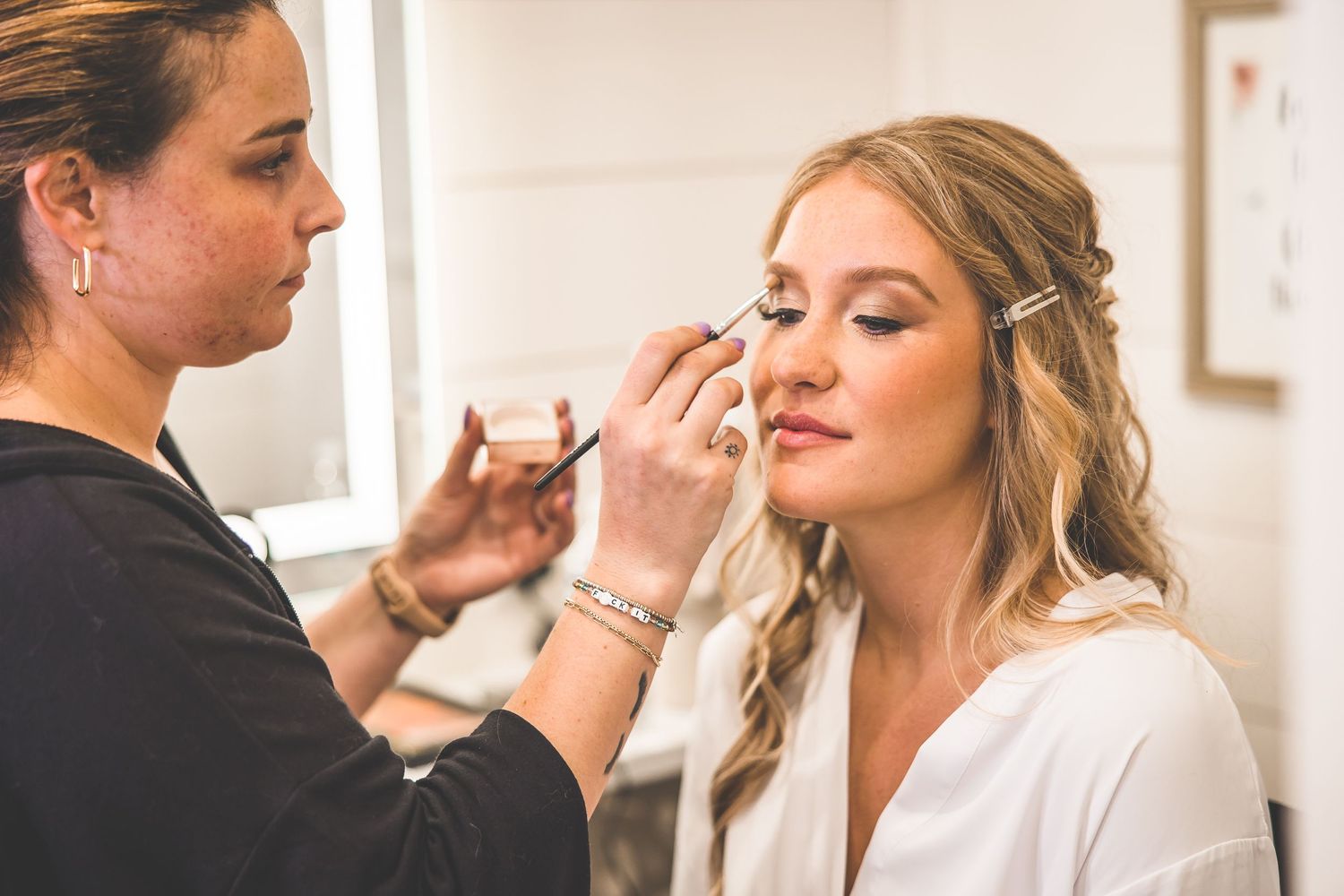 A makeup artist applies cosmetics to a client wearing a white robe during wedding preparations.