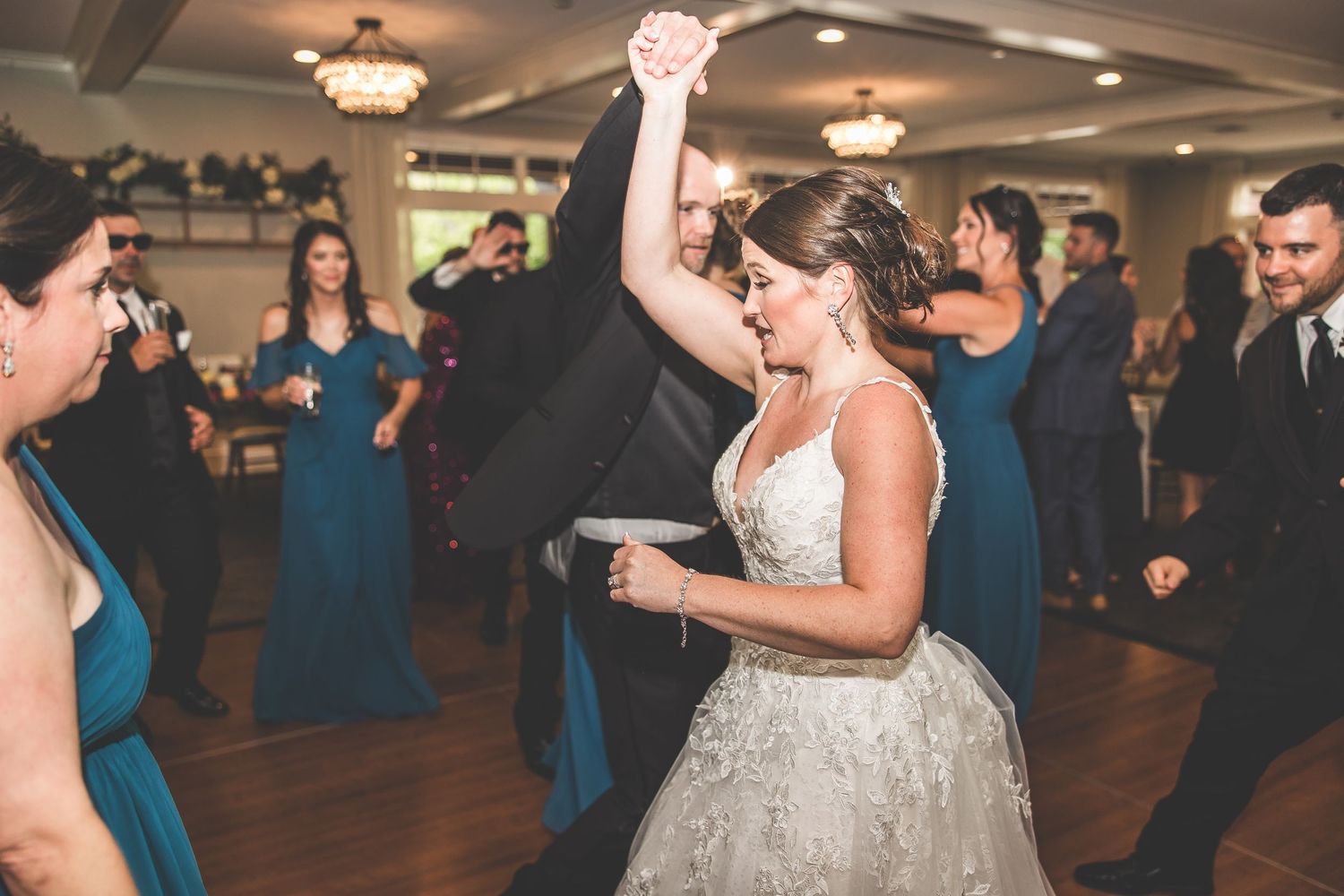 Wedding guests and couple dancing together on a dark hardwood dance floor under chandeliers.