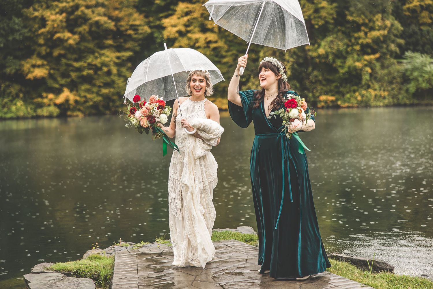 Wedding party with umbrellas poses by a lake on a cloudy autumn day.