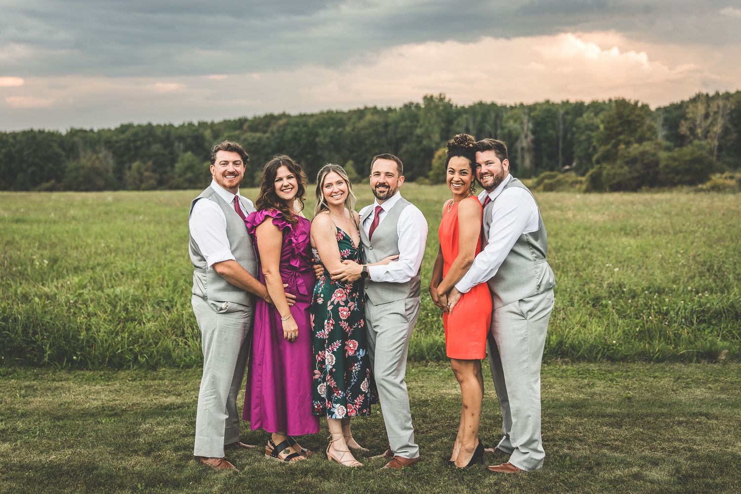 Wedding party poses in field at sunset wearing gray suits and burgundy dresses.