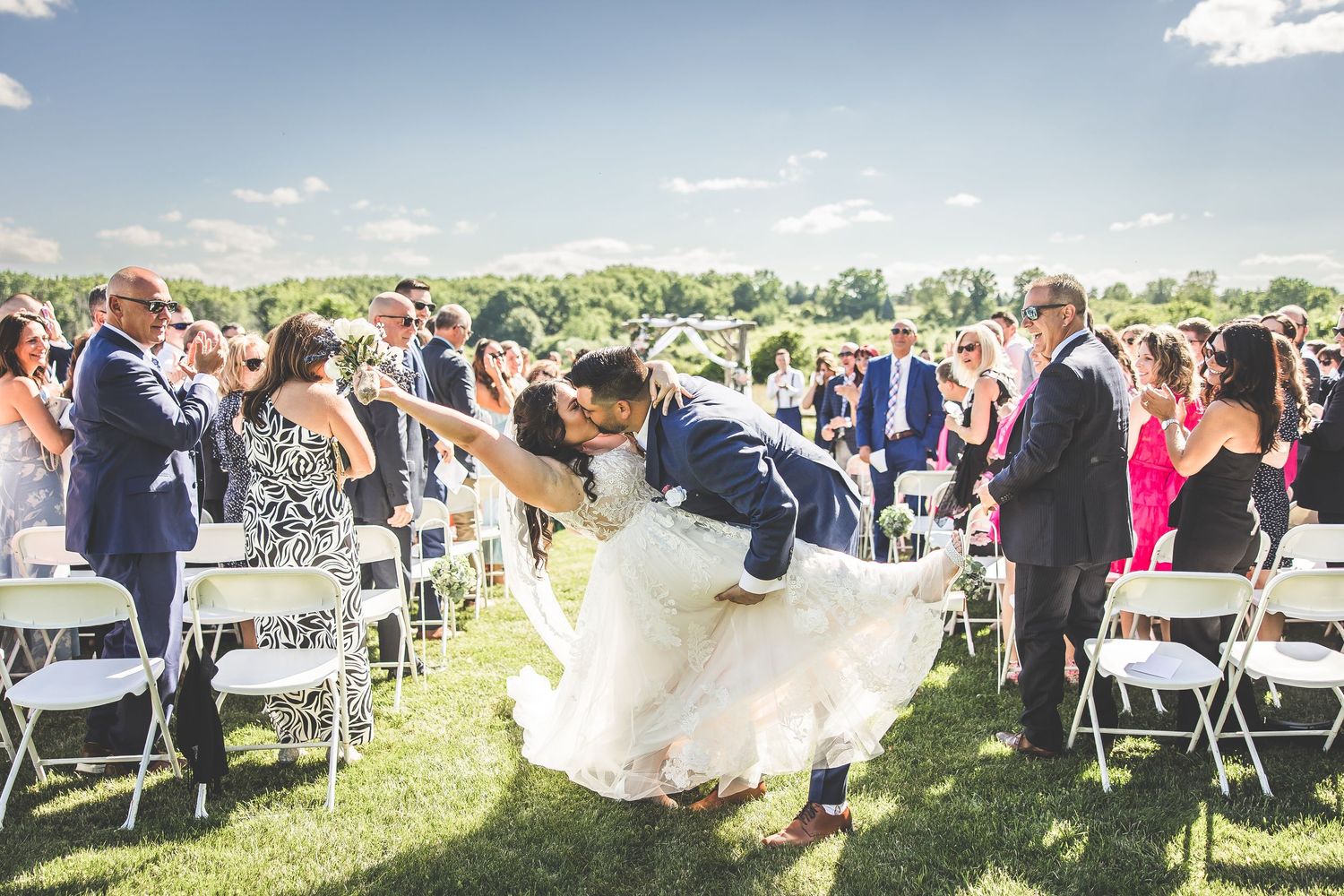 Wedding guests seated in white chairs on a sunny outdoor lawn during ceremony.