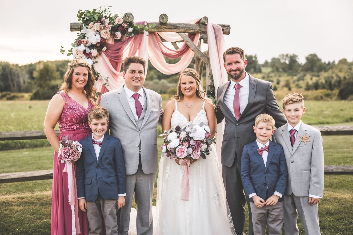 Wedding party in pink dresses and gray suits poses for formal photos in an outdoor setting.