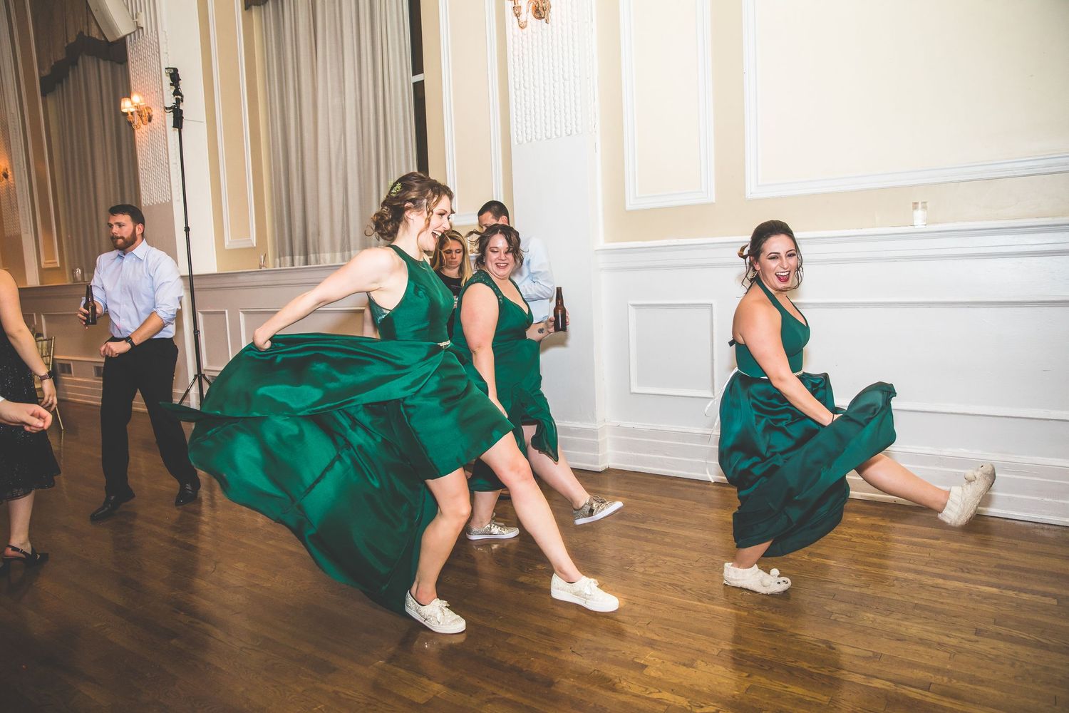 Wedding party in emerald green dresses dancing on a wooden floor during reception celebrations.