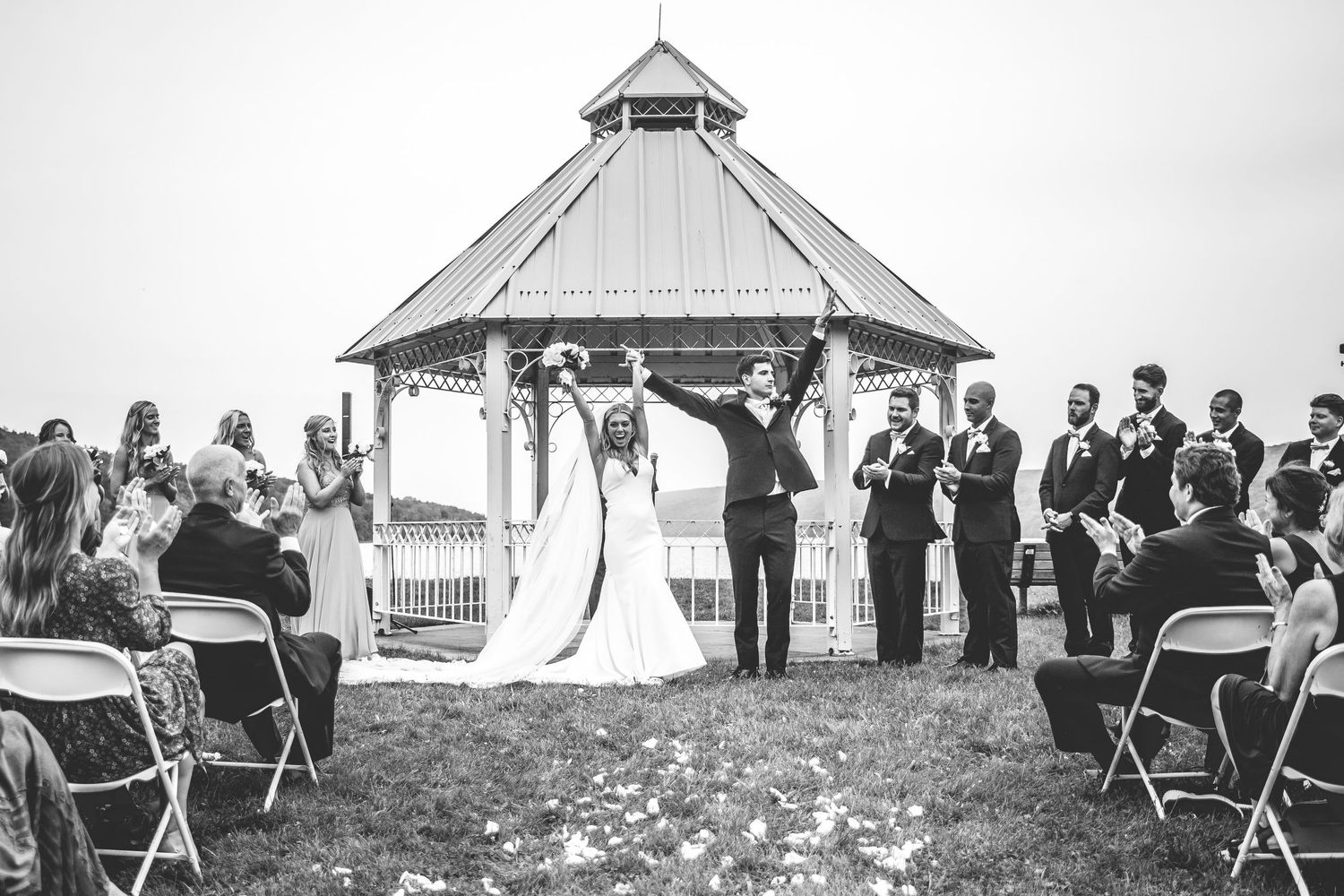 Wedding celebration panorama showing guests, white gazebo, and scattered flower petals on grass.