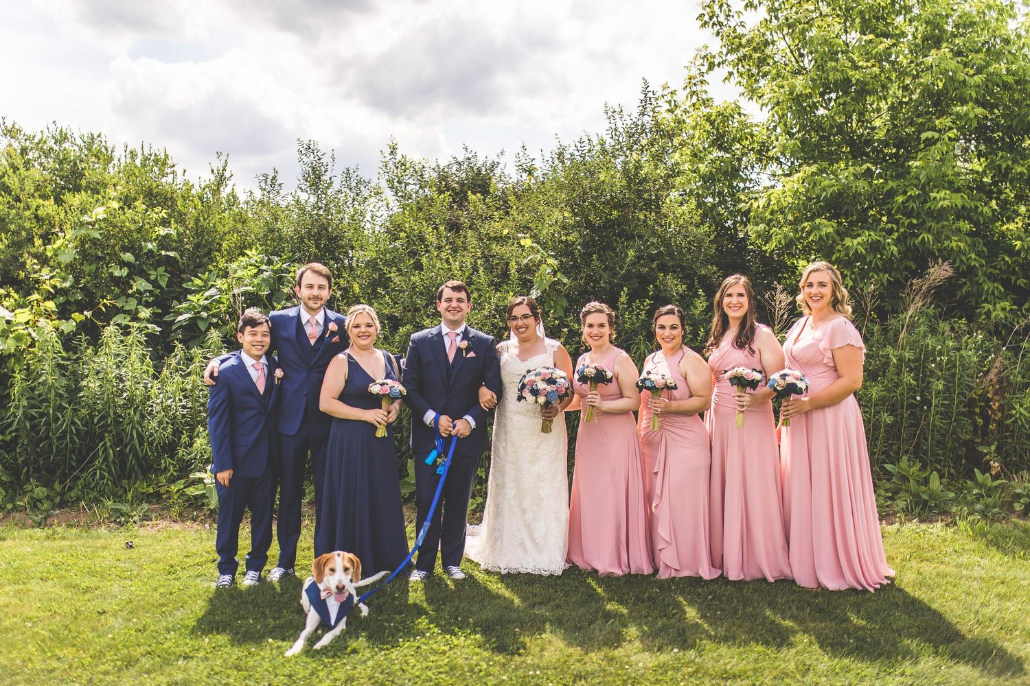 Wedding party members pose together on a grassy hill with trees in the background.